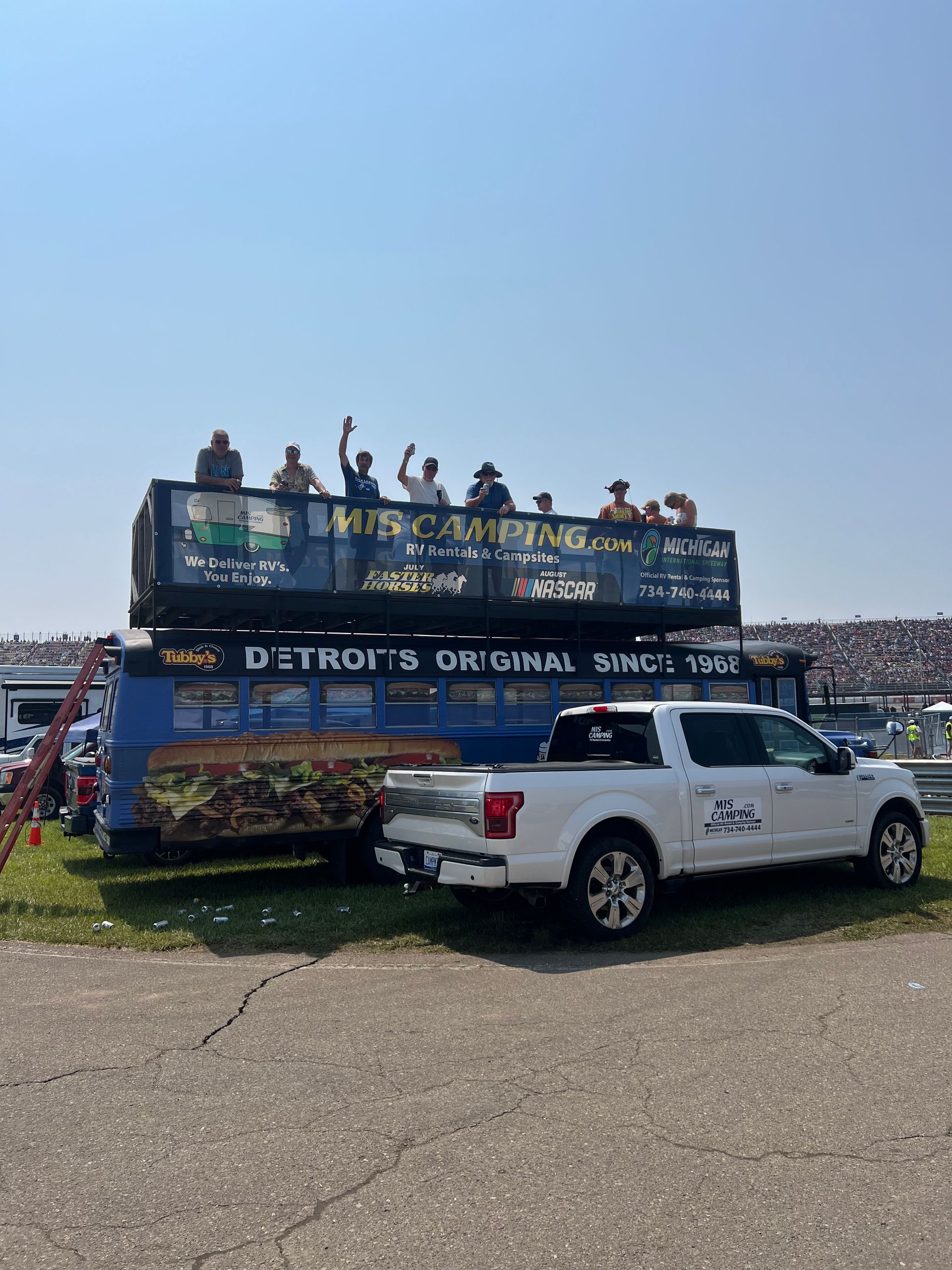 Two-story food truck with people waving on top, parked next to a white pickup truck.
