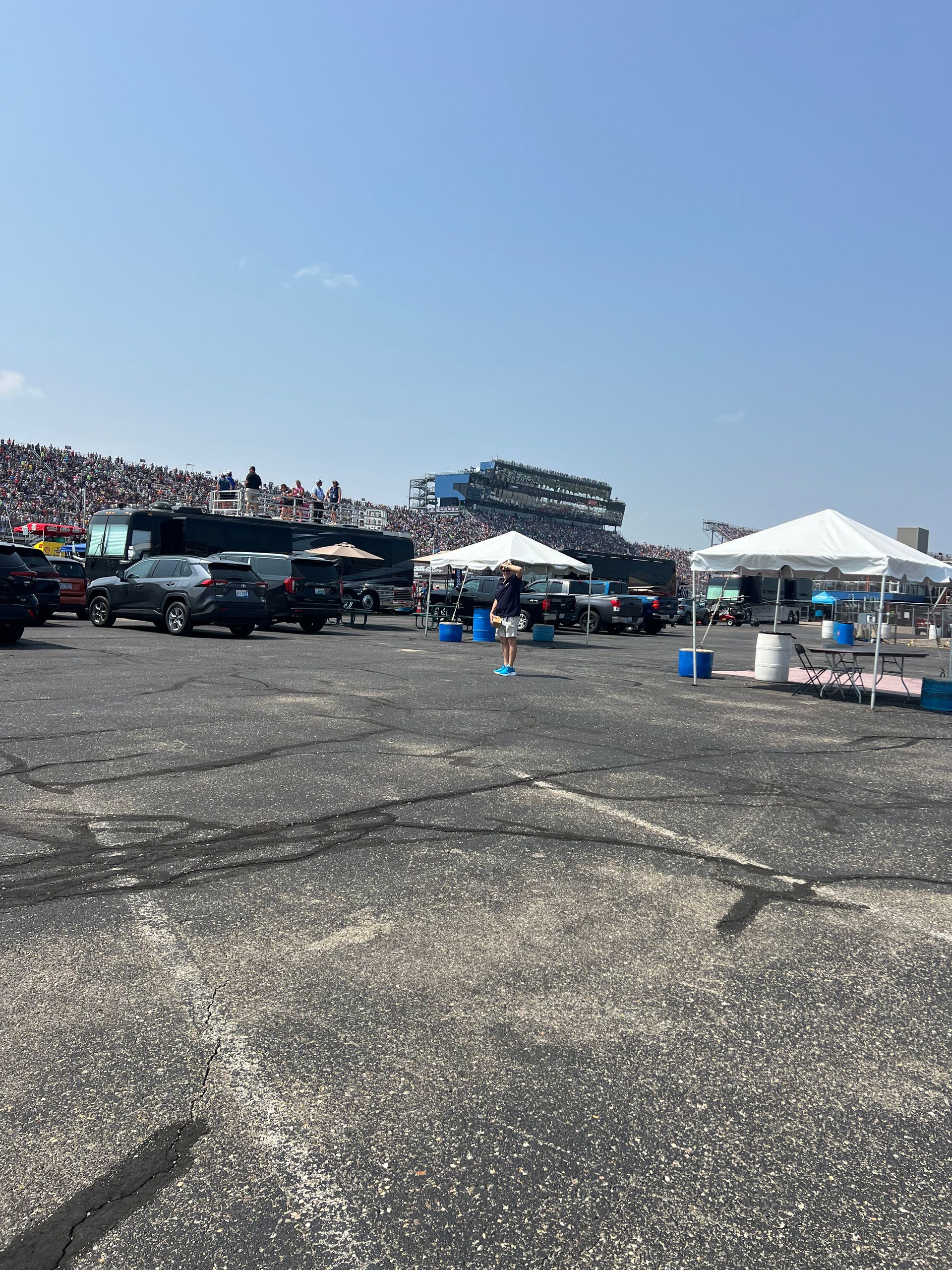 Large crowd at an outdoor event with race cars and tents, under a clear blue sky.