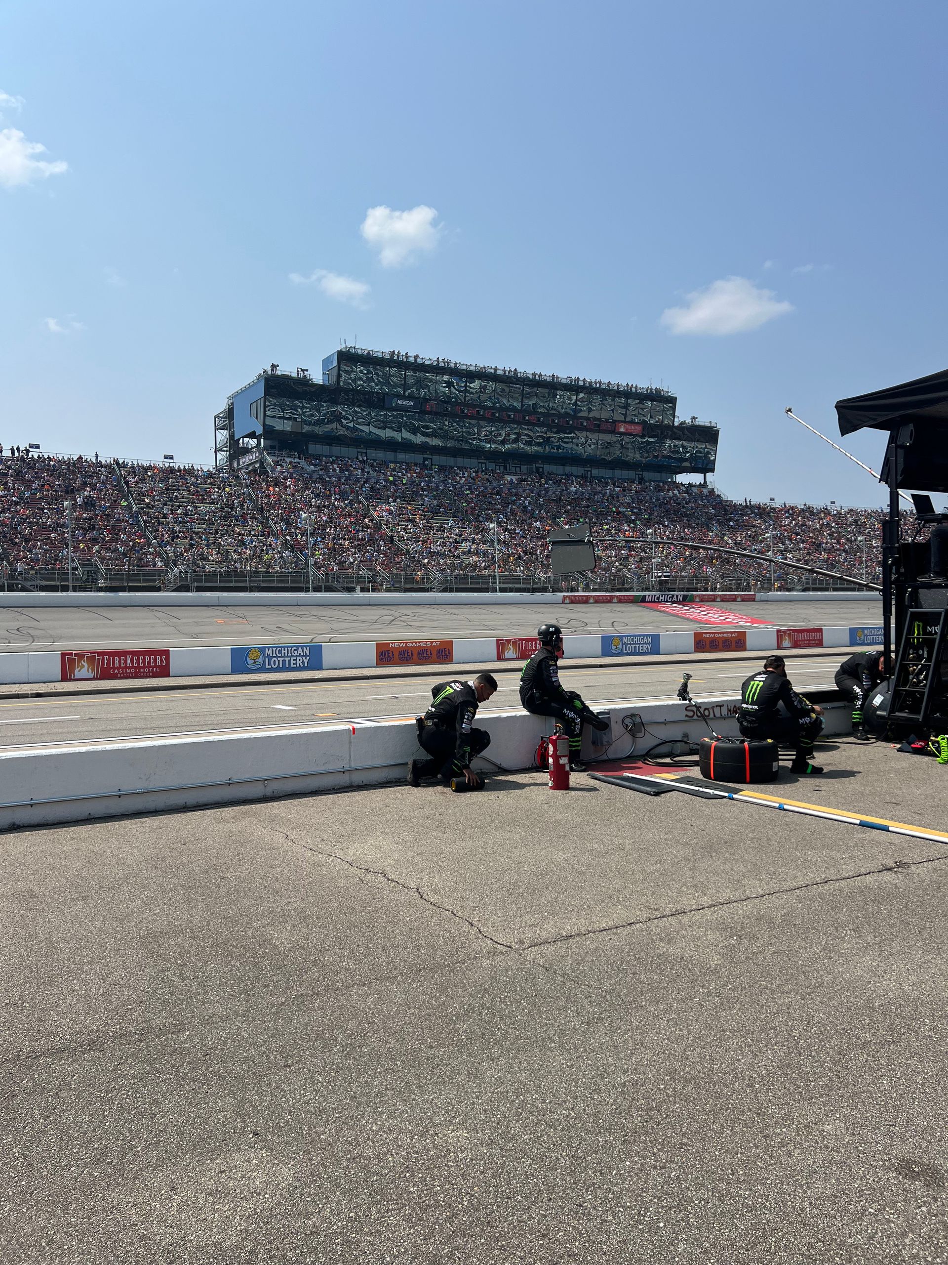 Race track pit crew working during a race. Bleachers filled with spectators under a bright blue sky.