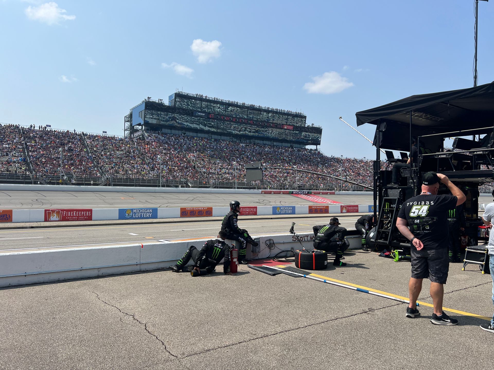 Race car pit crew at work on a racetrack, large crowd in the background. Bright sunny day.