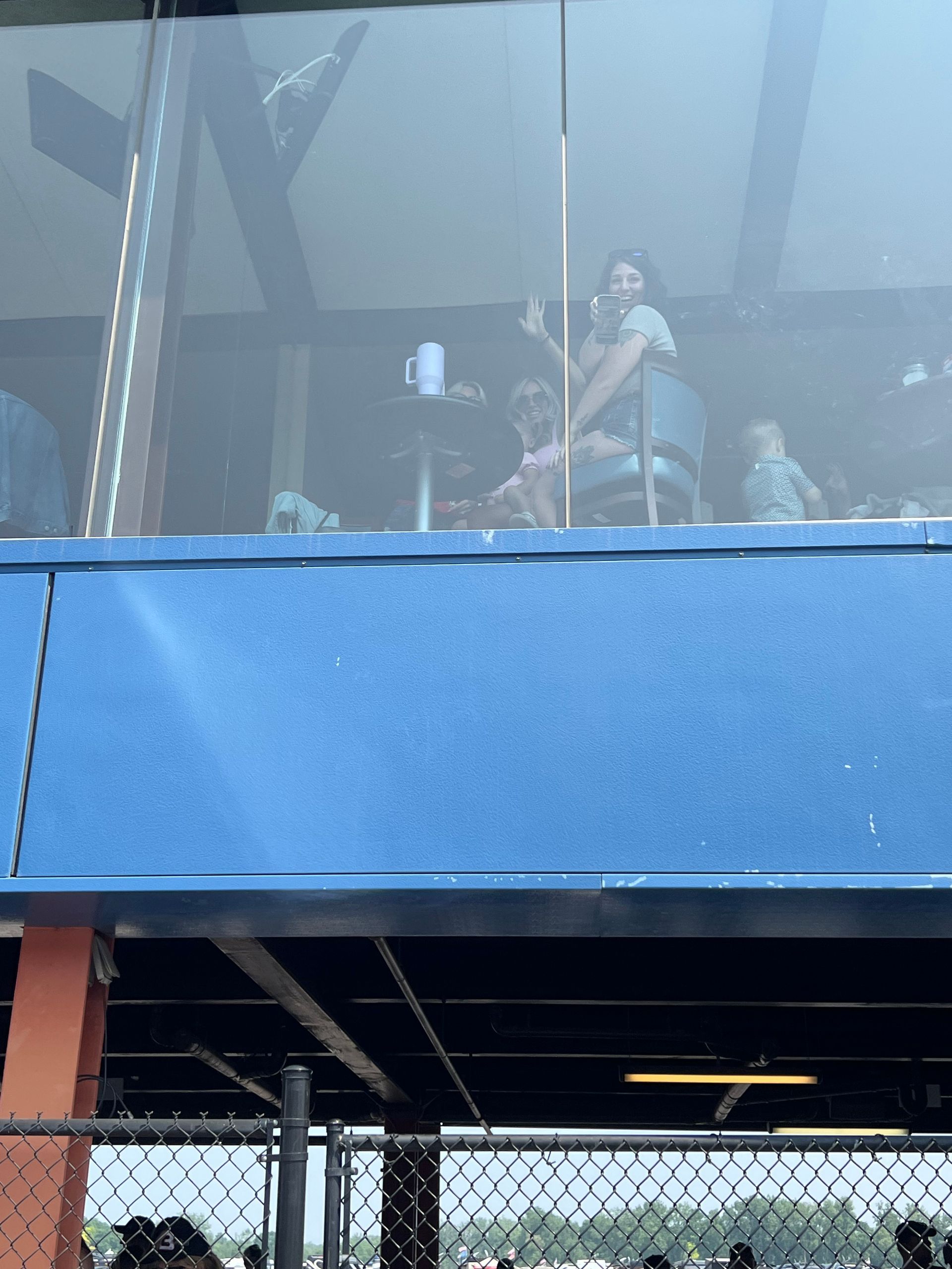 People waving from a box above a baseball field. Blue wall, glass windows, black fence.