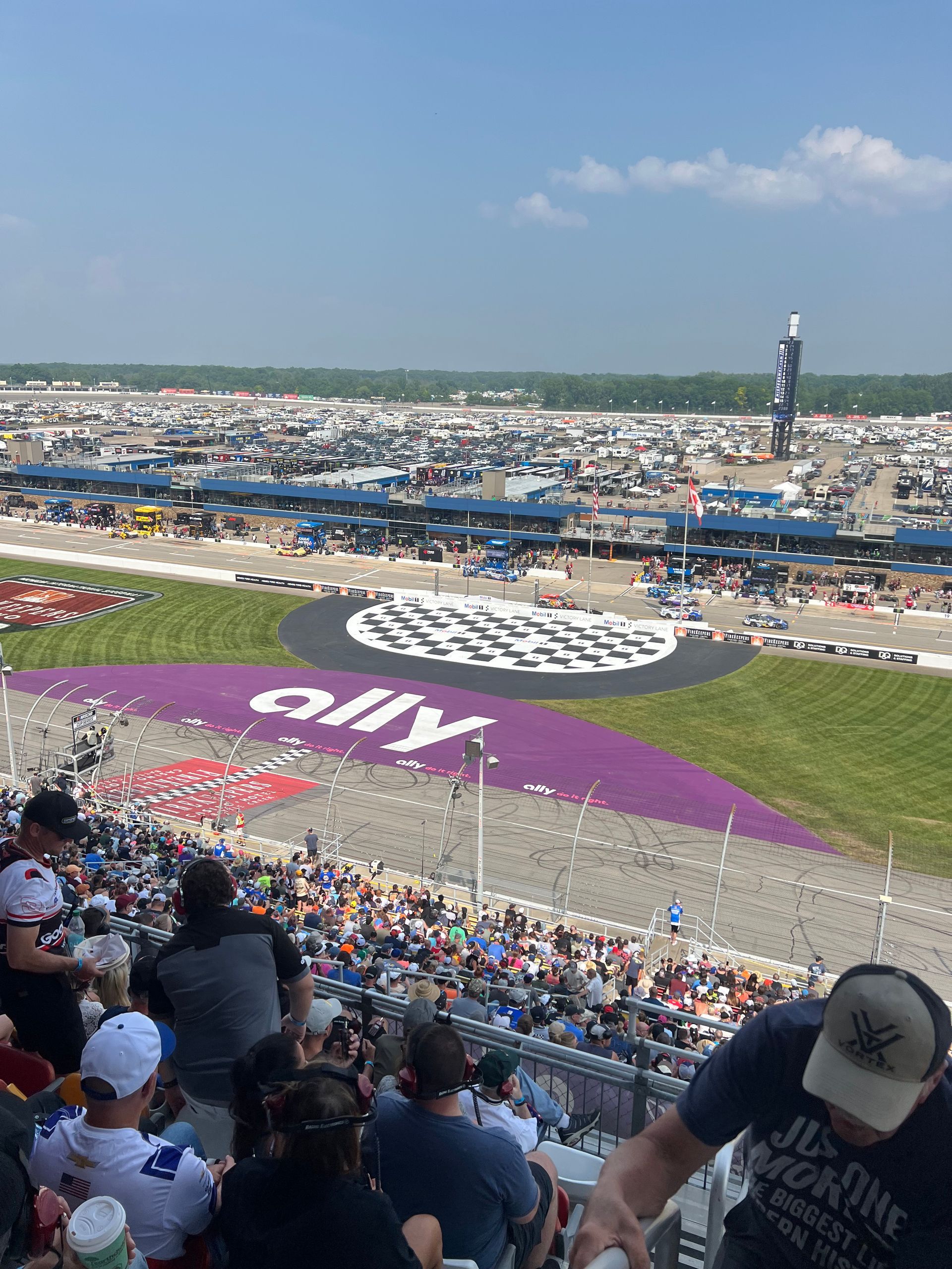 View of a race track with fans in the stands, Ally logo in infield, and cars on the track.
