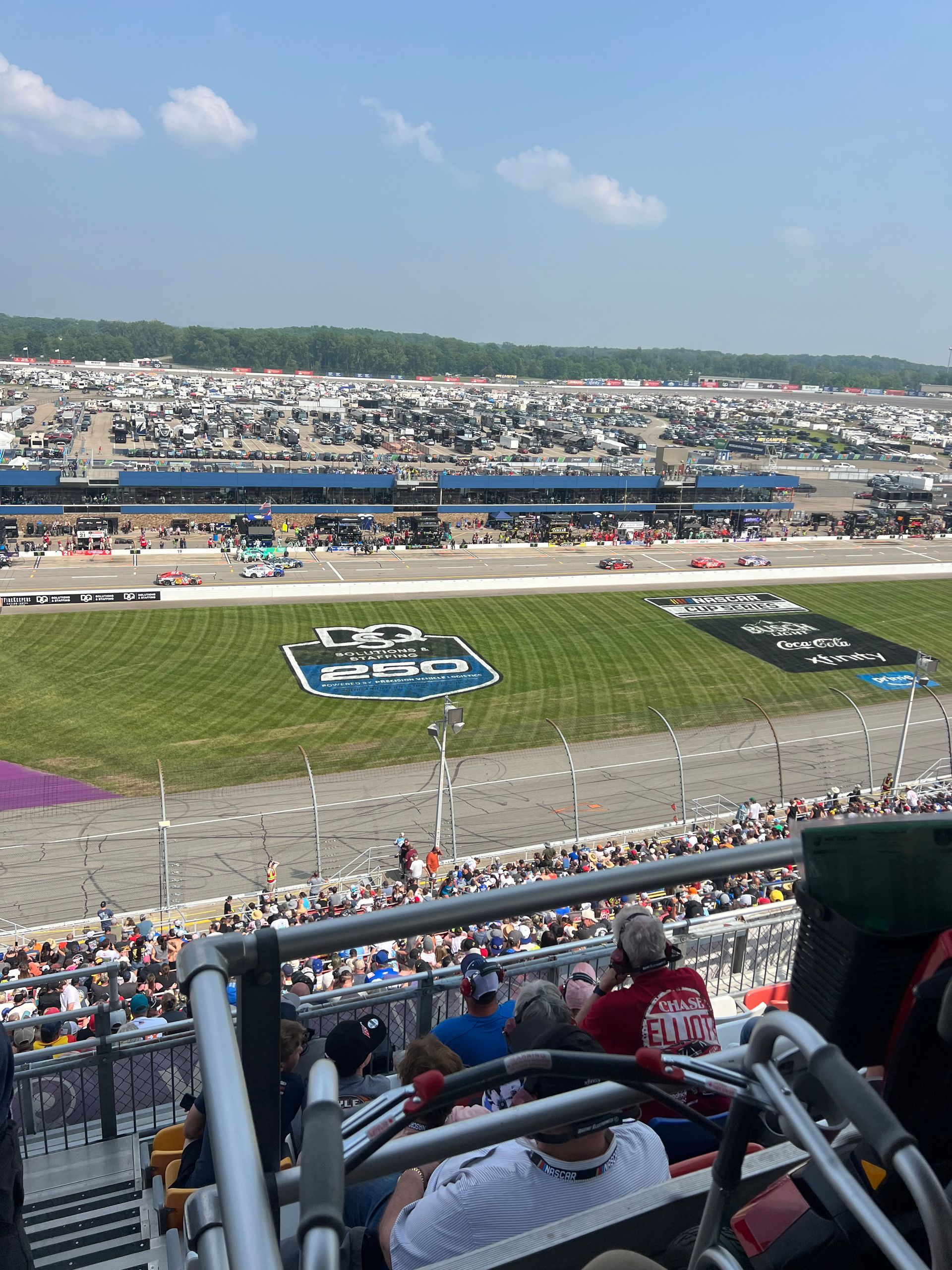 View from stadium seats overlooking a racetrack and parking lot; sunny, blue sky.