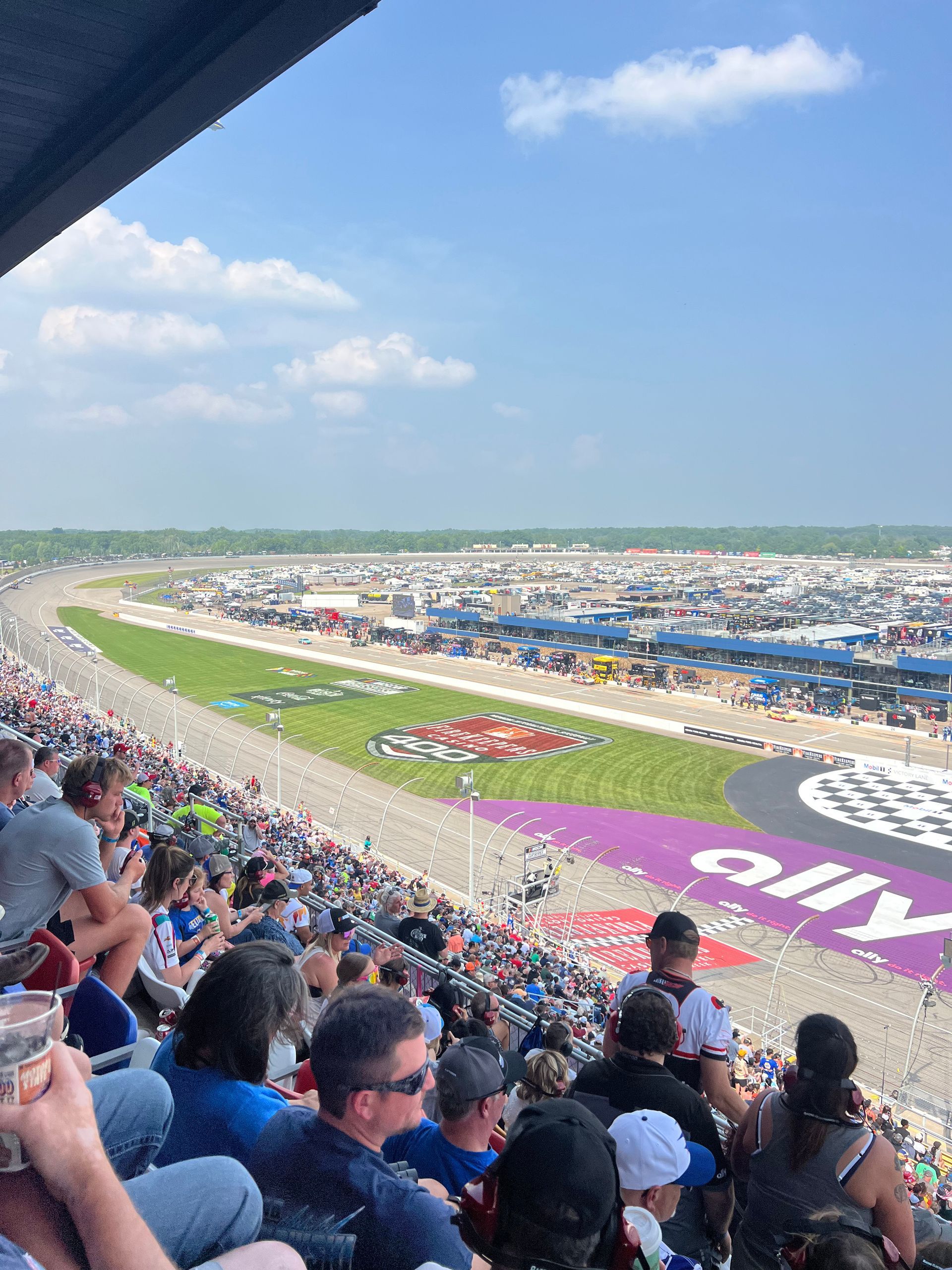 Spectators watch a NASCAR race from the stands at a speedway on a sunny day.