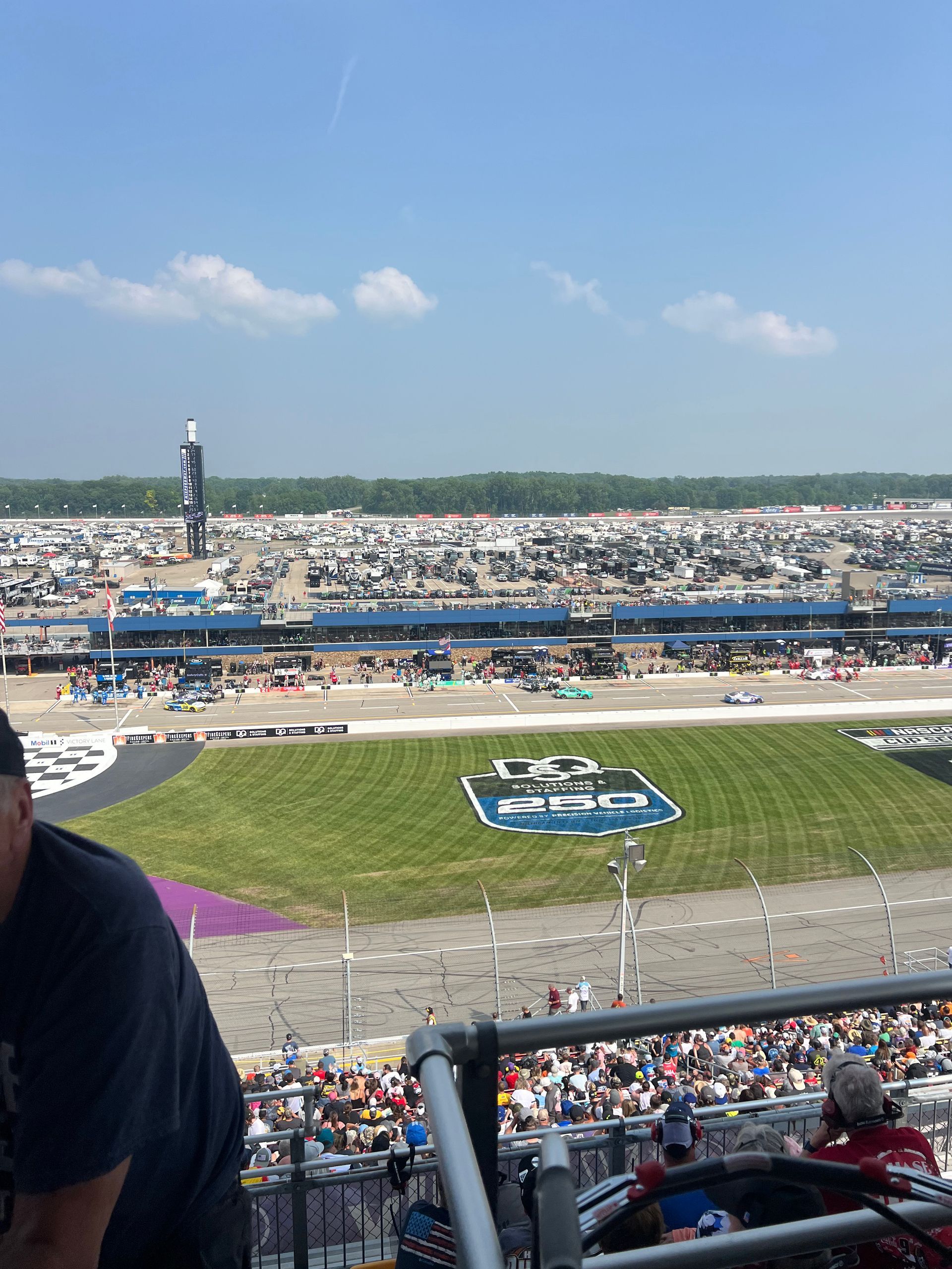 Race track with a large logo on the infield grass, stands filled with people, and cars on the track.