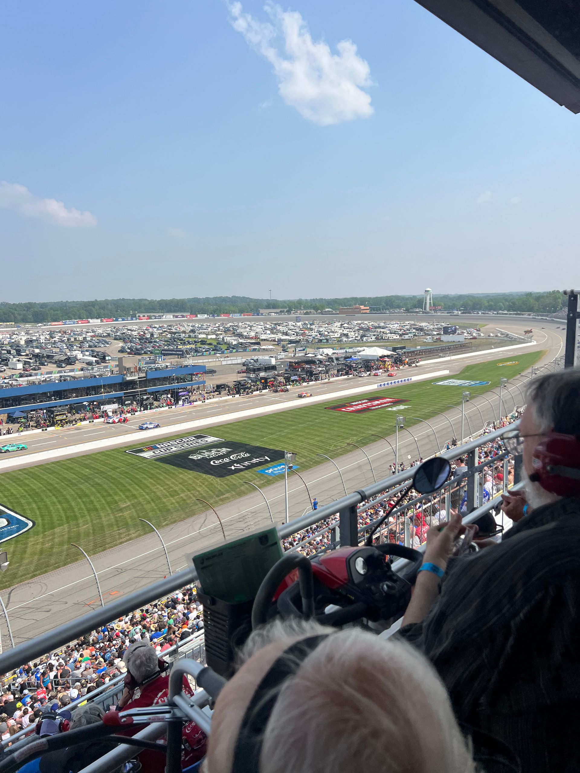 Race track scene with grandstand, spectators, and racing cars, sunny day.