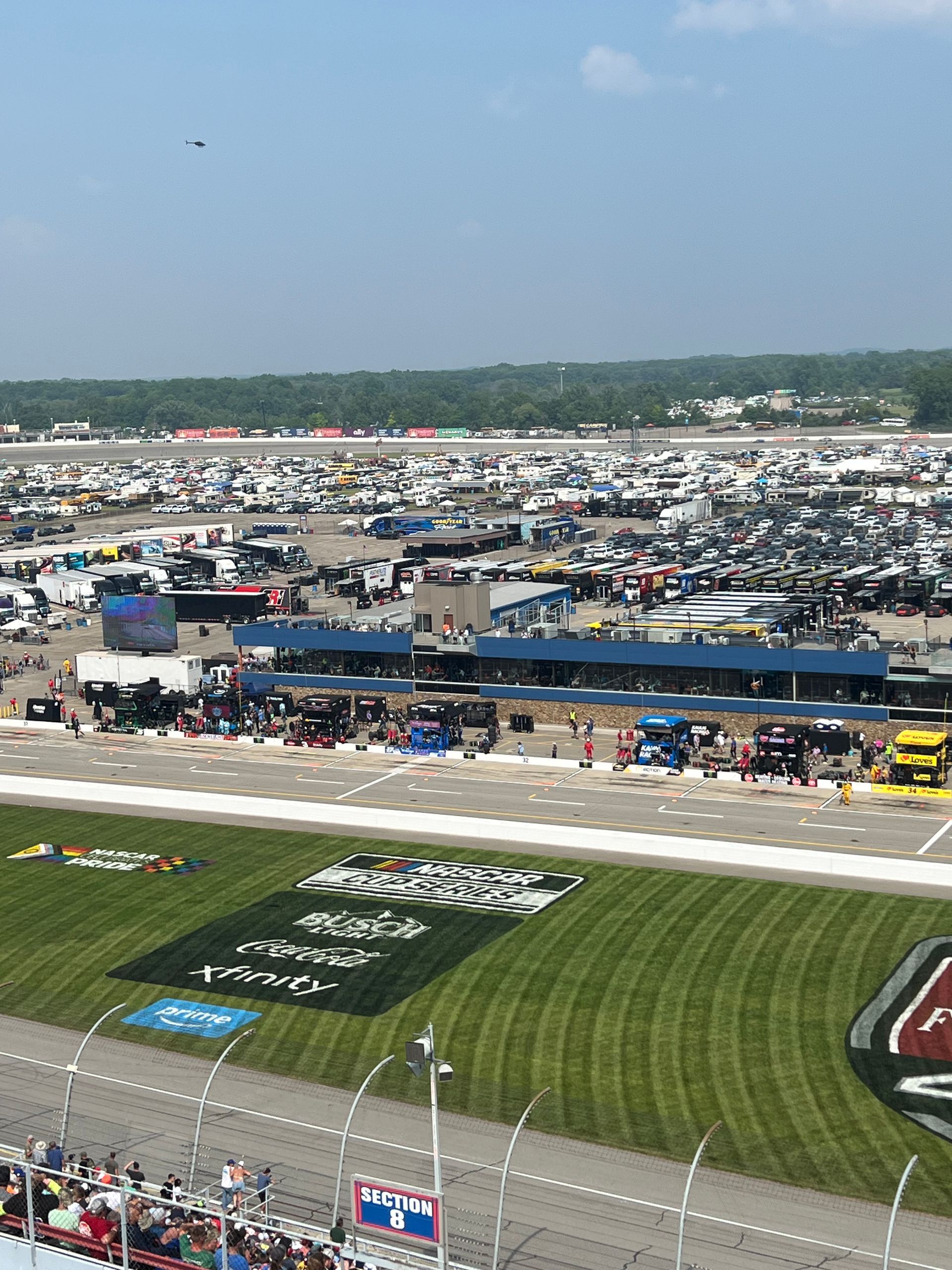 Race track scene with cars, pit crews, and grandstands under a bright sky.
