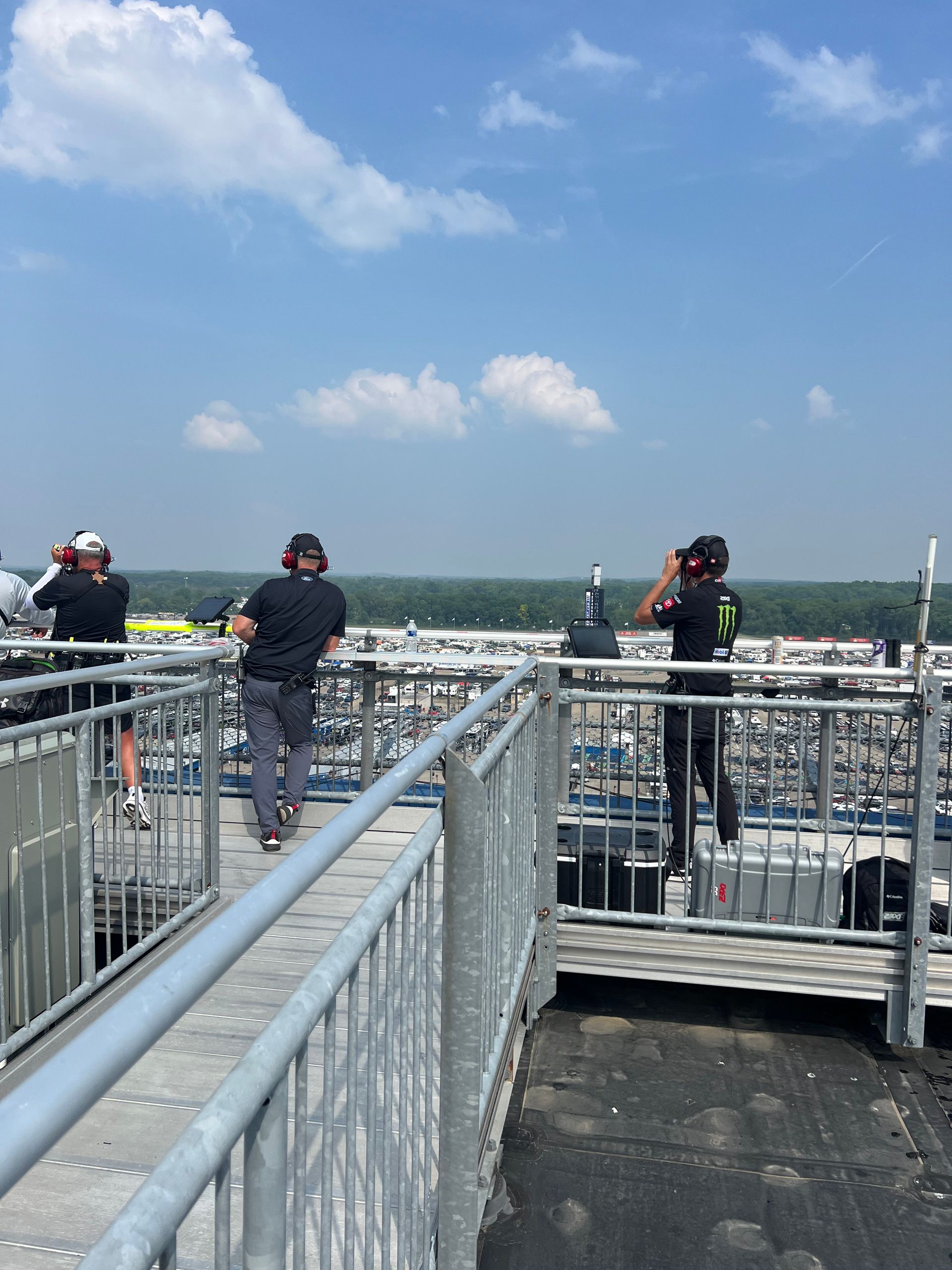 Three people overlooking a race track from a high platform, using binoculars under a blue sky.