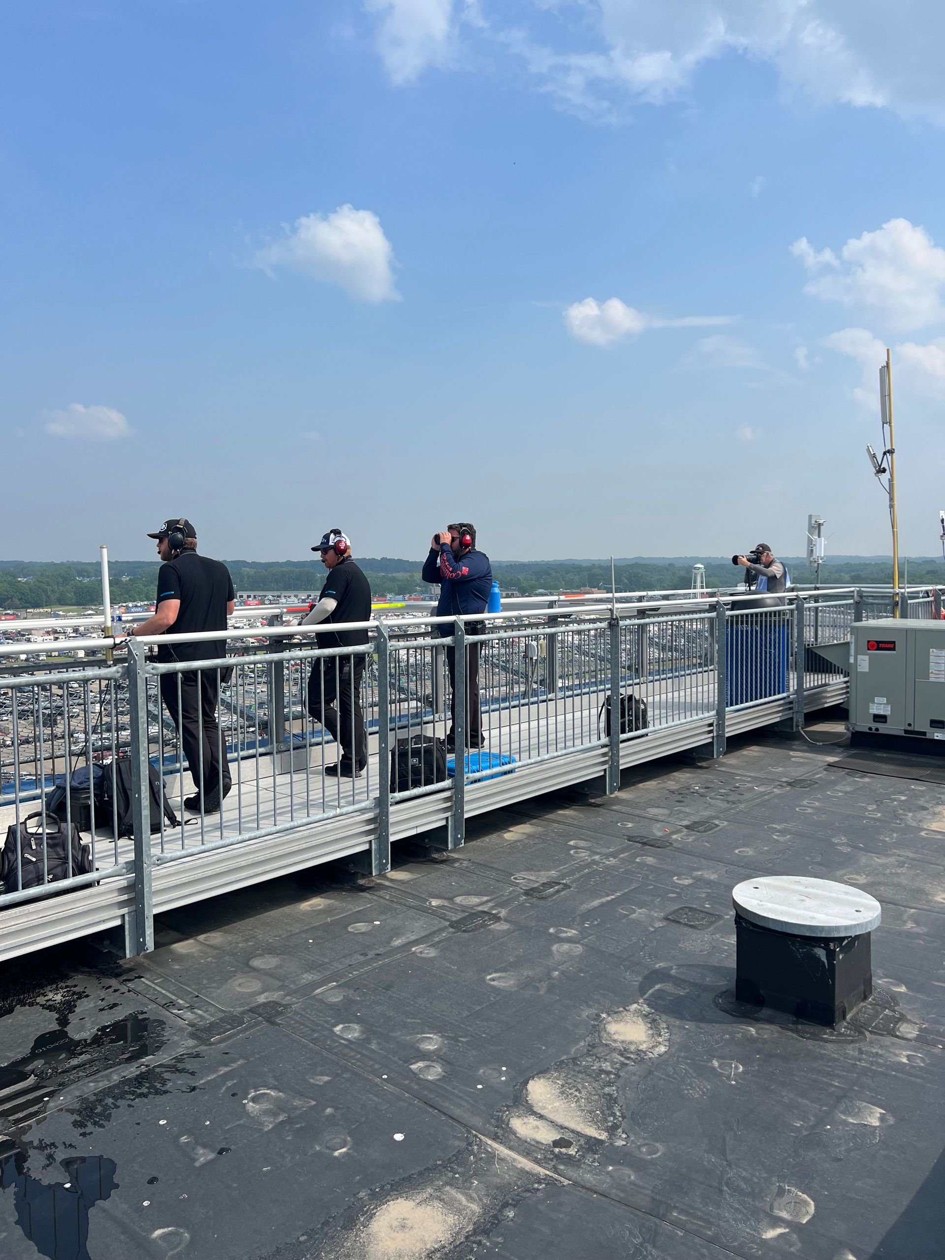 People on a rooftop walkway looking at something, with a city in the distance, under a blue sky.