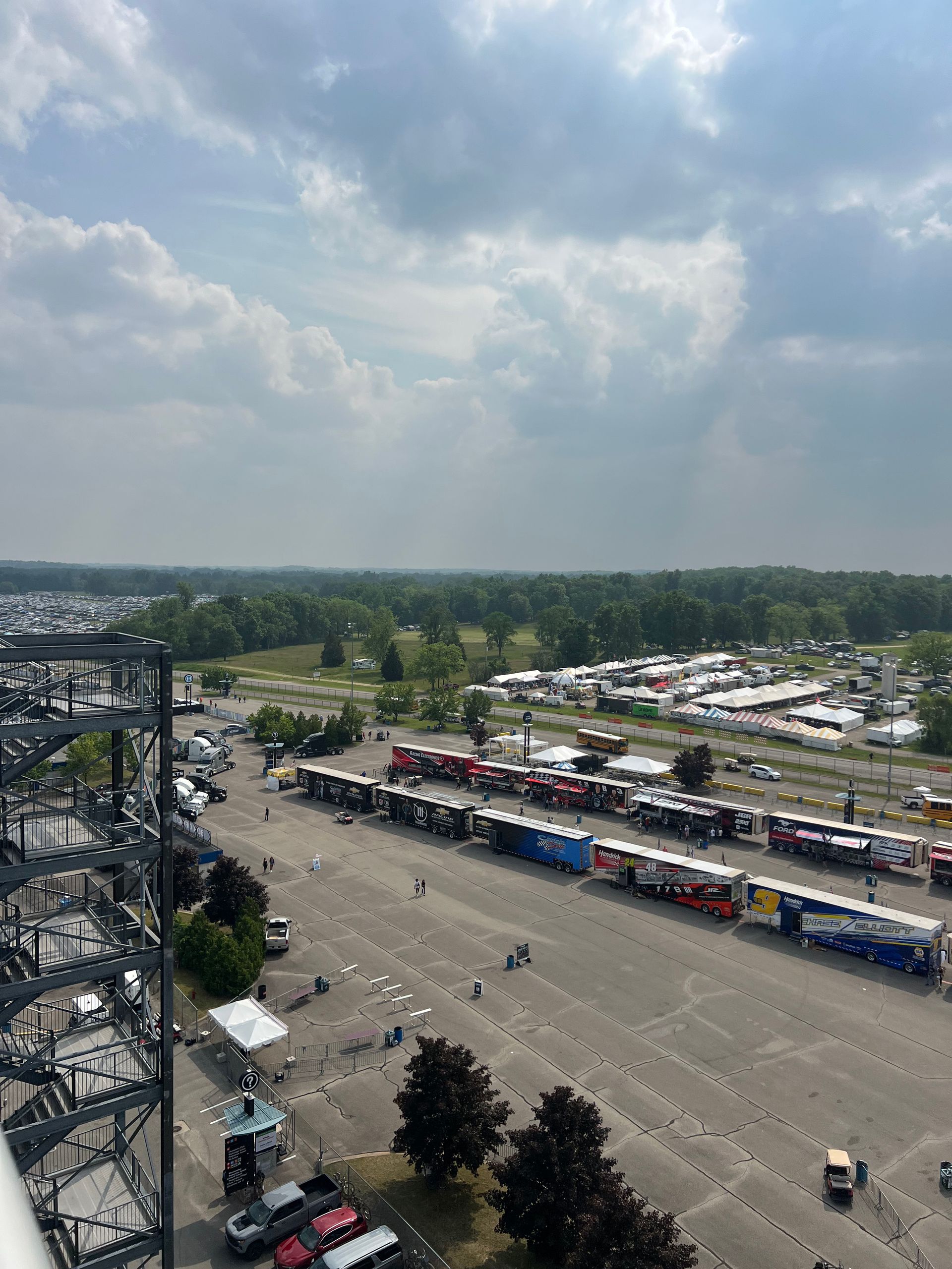 View from above of a racetrack pit area with semi-trucks, cars, and people. Blue sky with clouds.