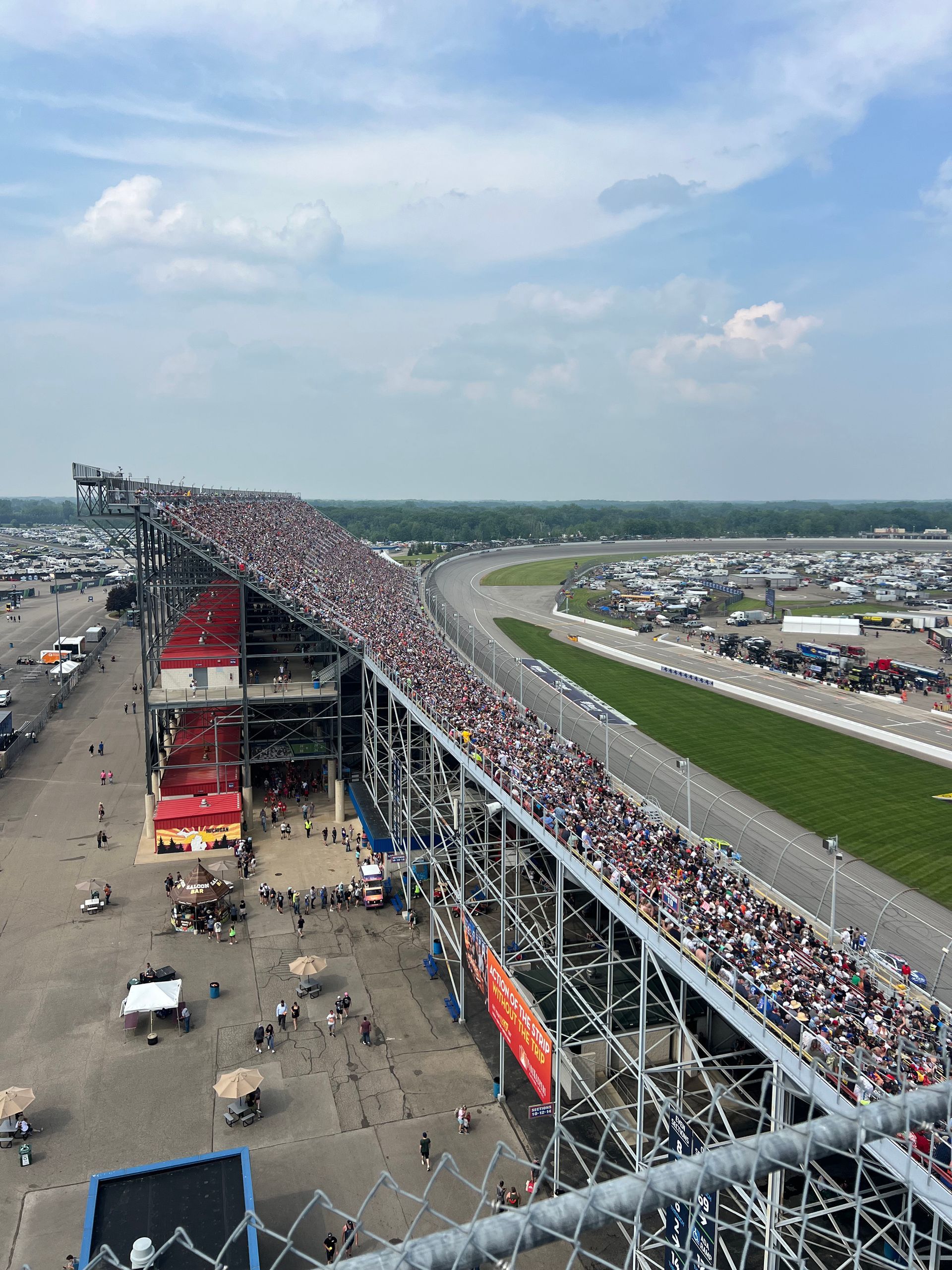 Race track grandstand packed with spectators, looking towards a curved track. Blue sky with clouds.