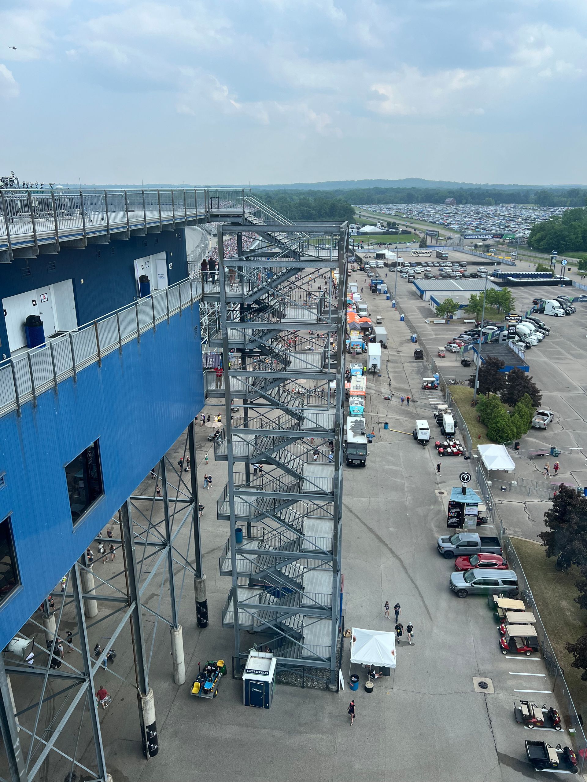 View from above of a racetrack and parking lot; blue structure, people, vehicles.