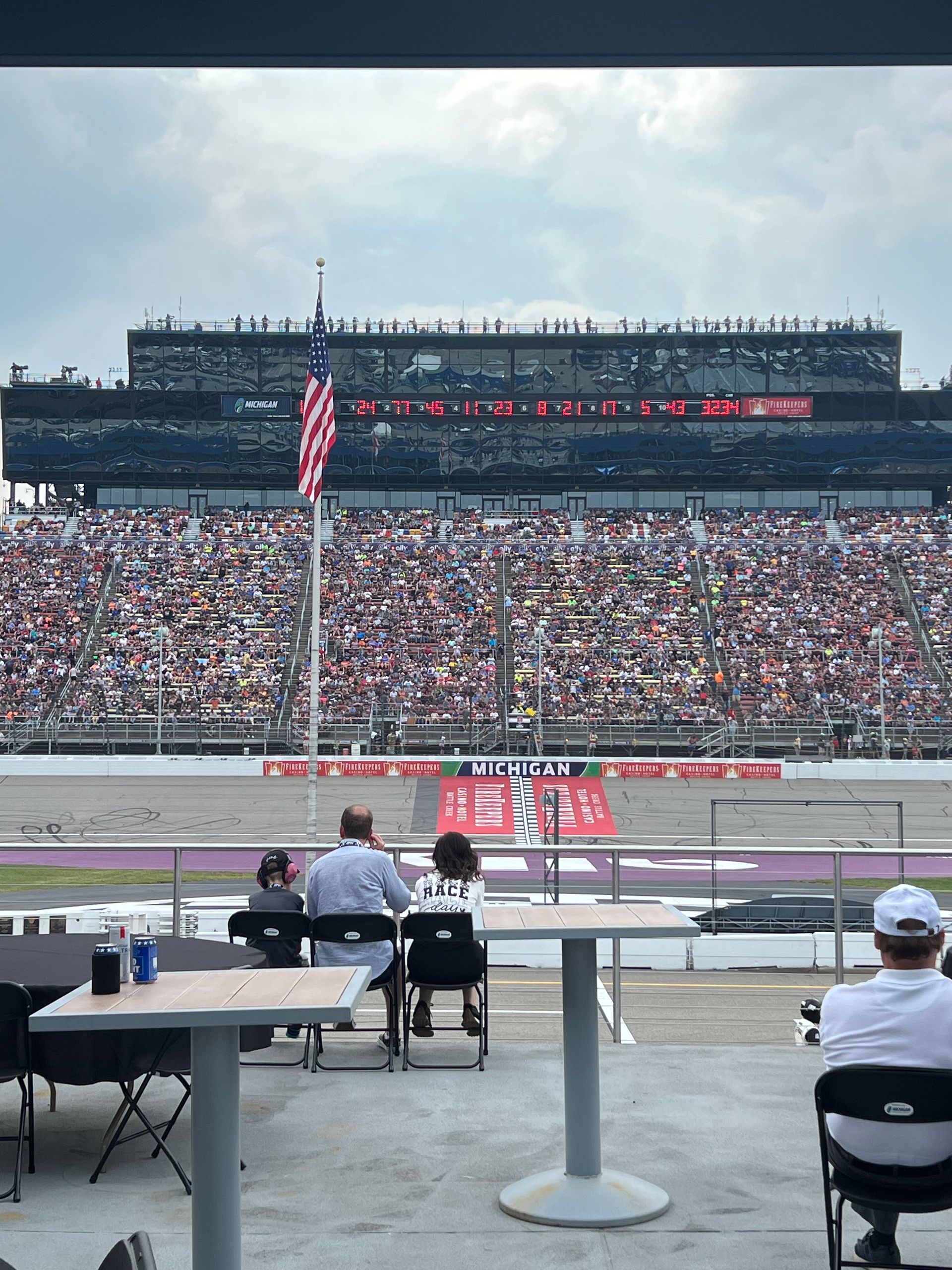 People watching a race from a stadium with an American flag. Crowds fill the stands.