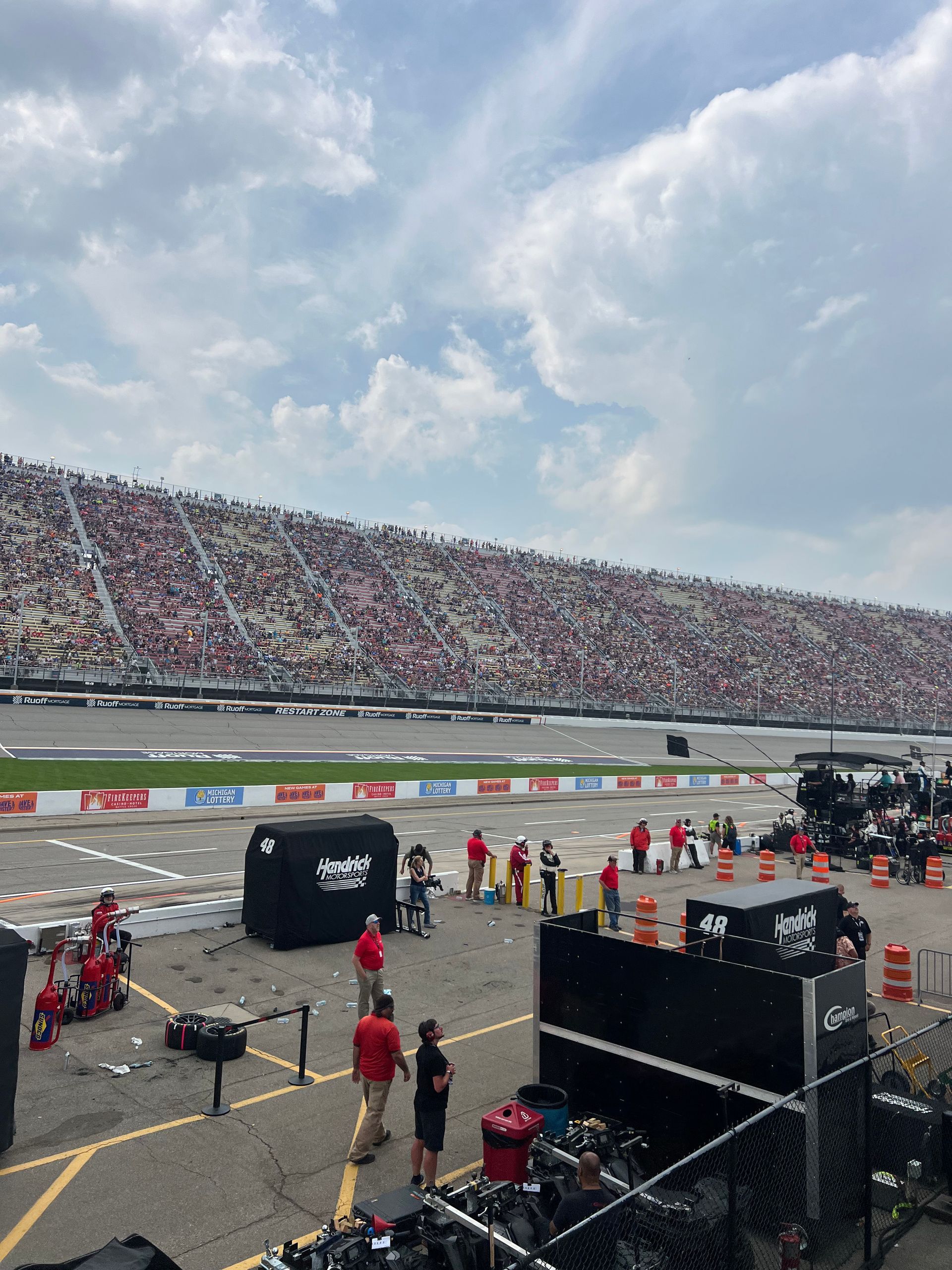 Race track scene with crowd in stands and crew members near pit lane under a cloudy sky.