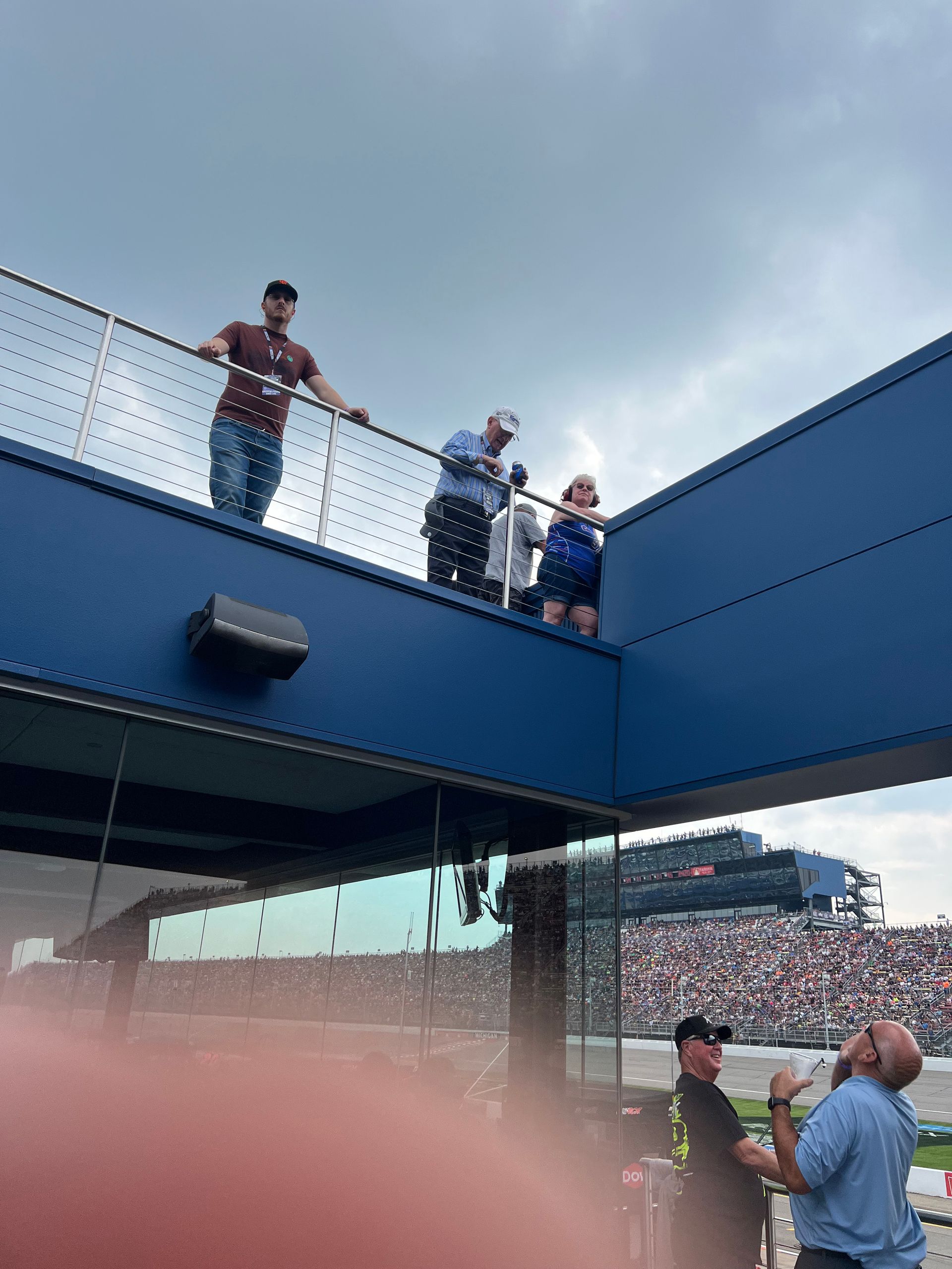 People watching a race from a blue elevated platform. Cloudy sky, stadium in the background.
