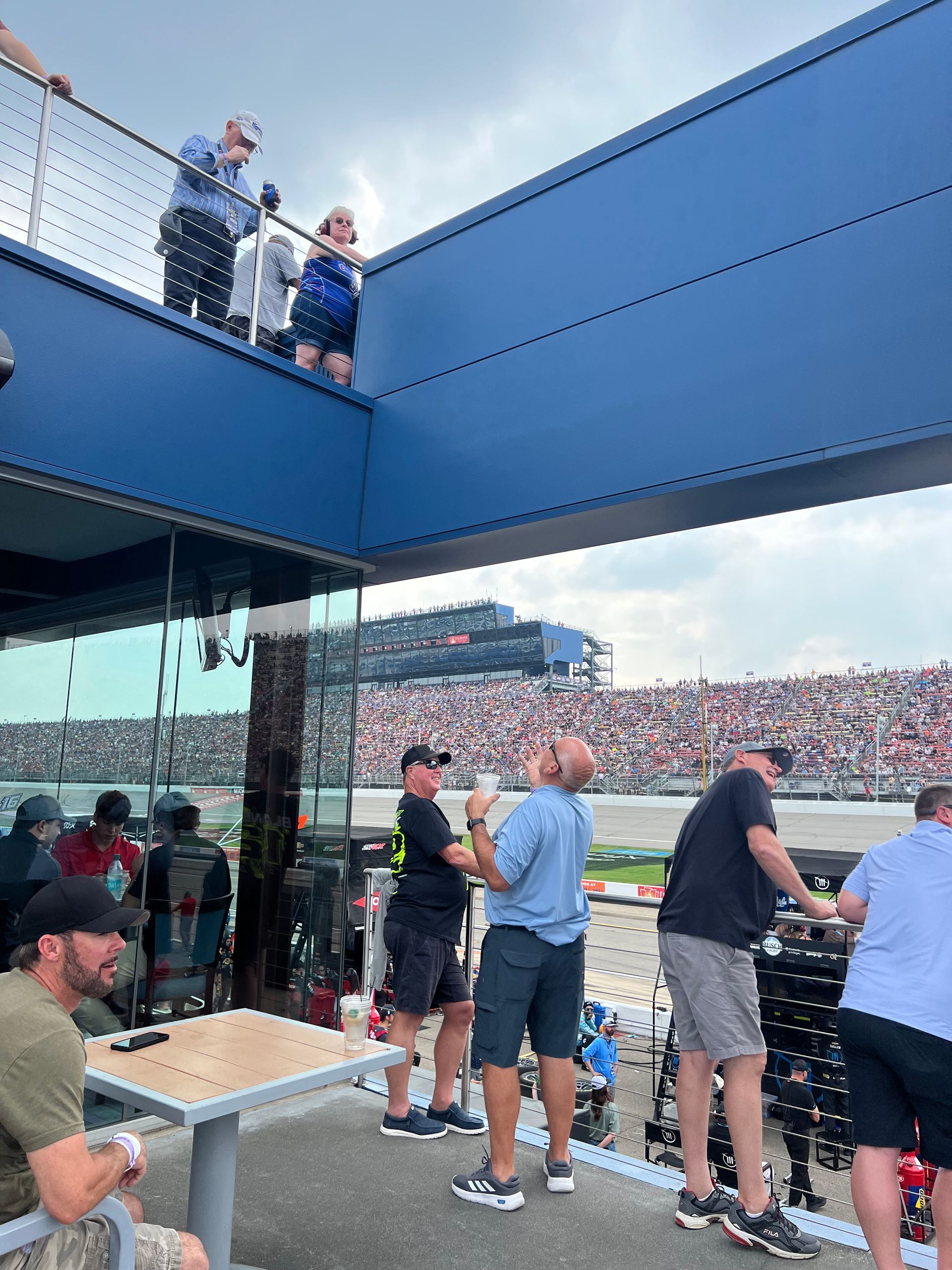People watch a race from a stadium viewing platform. The sky is overcast, and a crowd is in the distance.