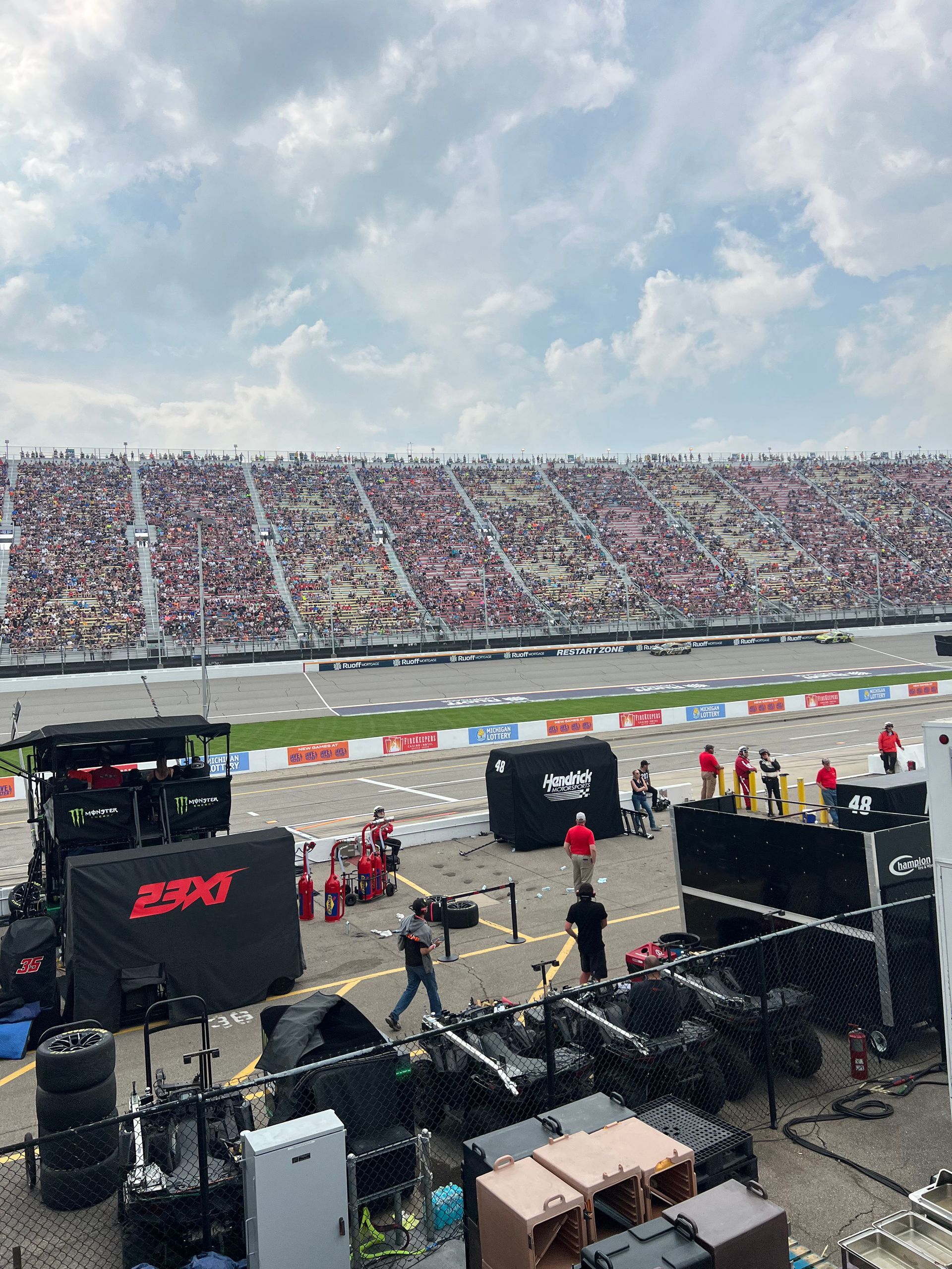 Race track scene with crowded stands under a cloudy sky. Crews and equipment are in the foreground.
