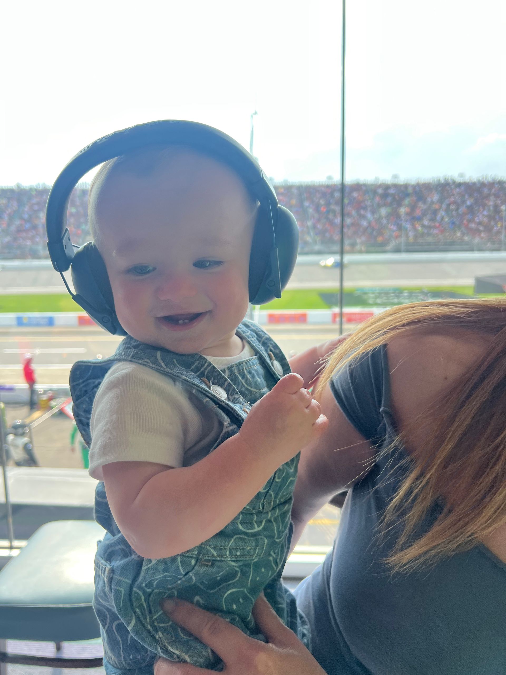 Baby wearing noise-canceling headphones smiles, held by a person with red hair, at a racetrack.