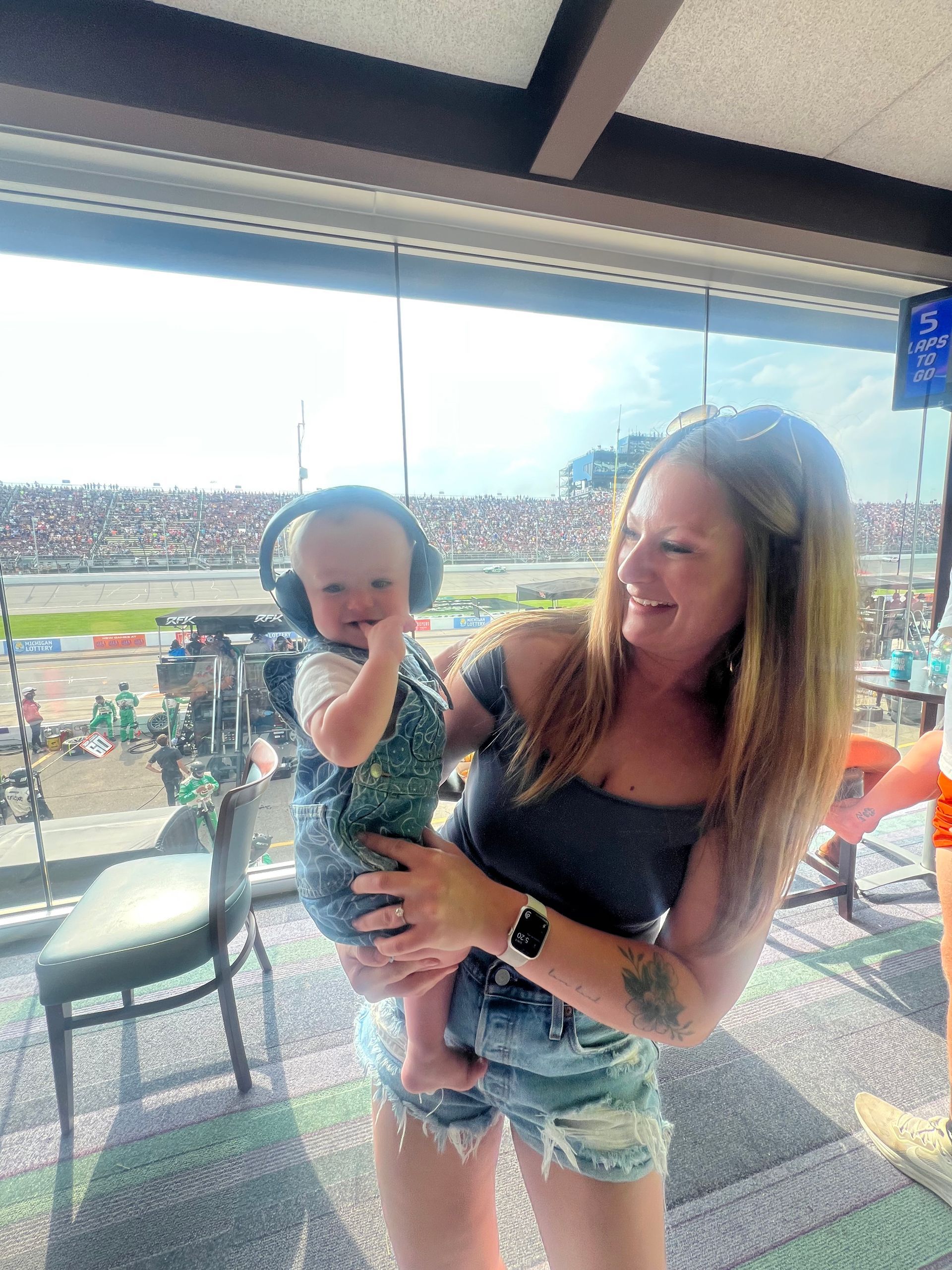Woman holding baby with noise-canceling headphones at a racetrack. Both smiling, in a spectator area.