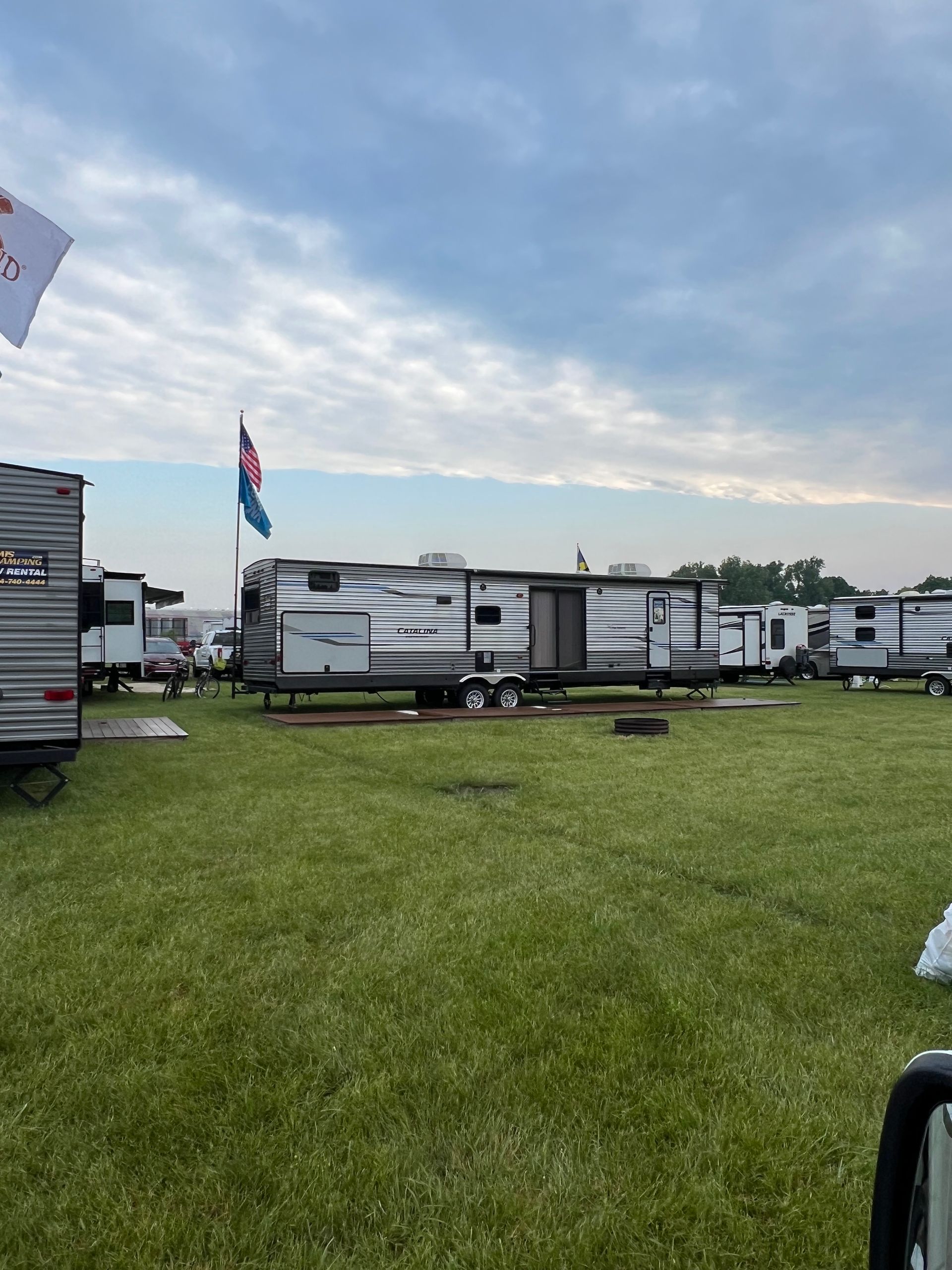 Campground with RVs parked on green grass under a cloudy sky. Flags visible.