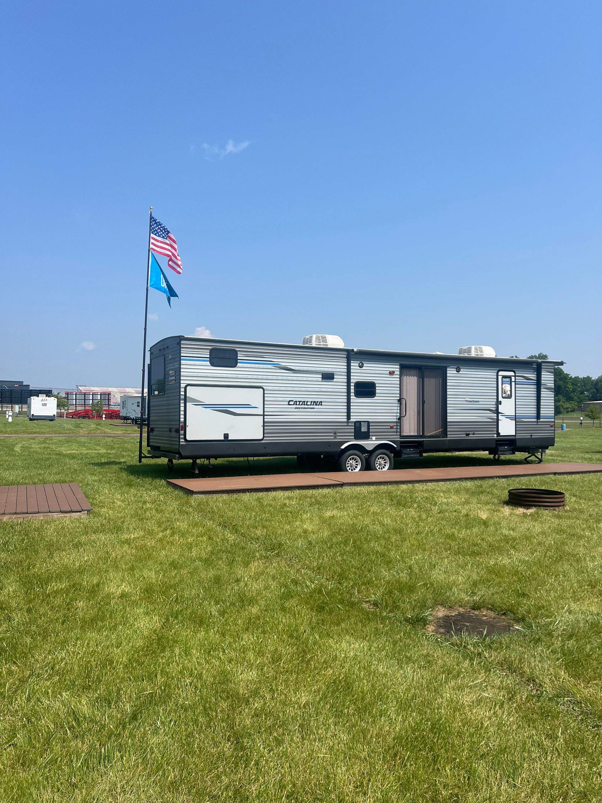 RV trailer with American flag and blue flag on a green field under a blue sky.