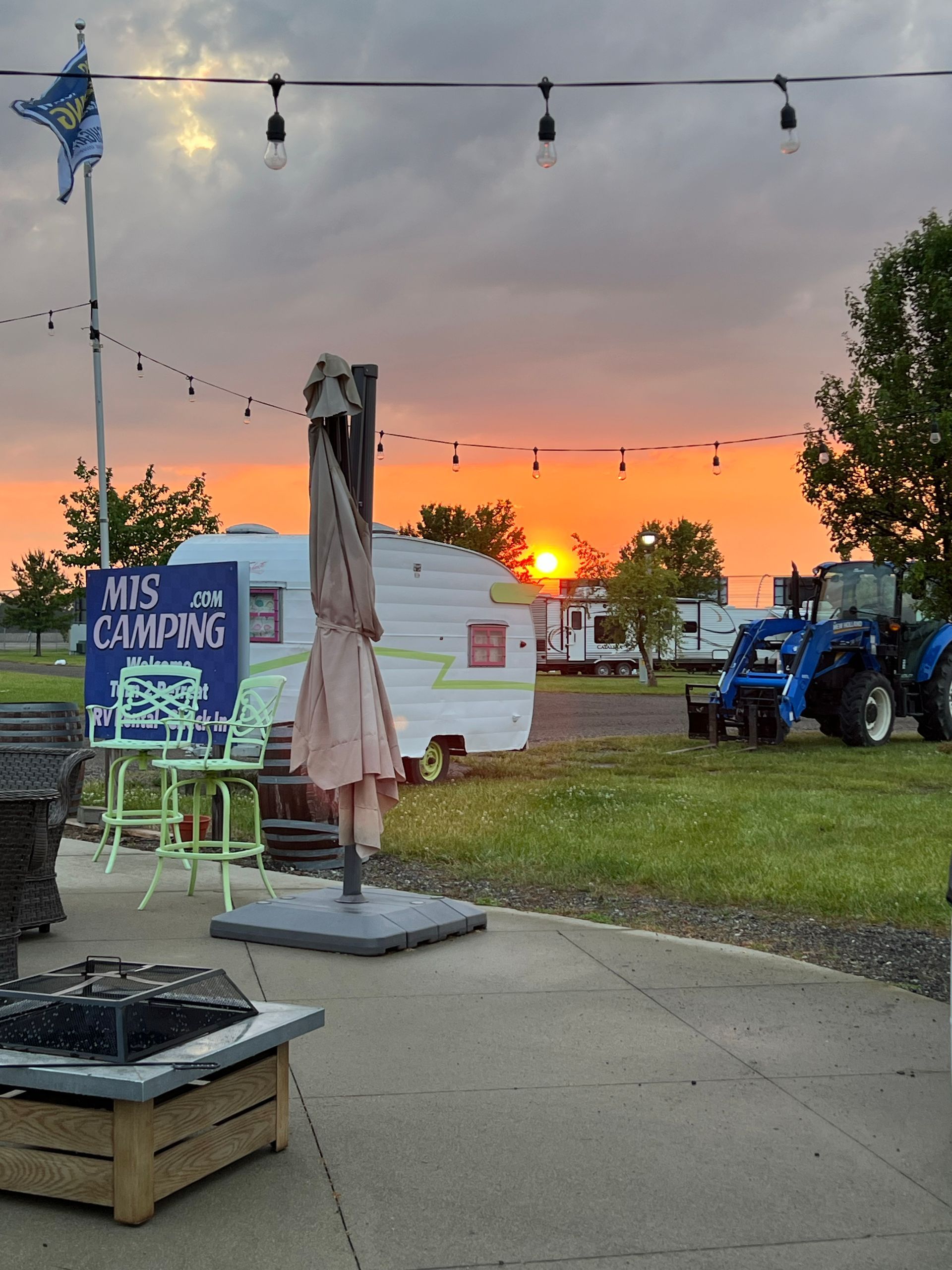 Sunset over a campsite with a vintage camper, picnic table, and tractor. String lights and a sign for 