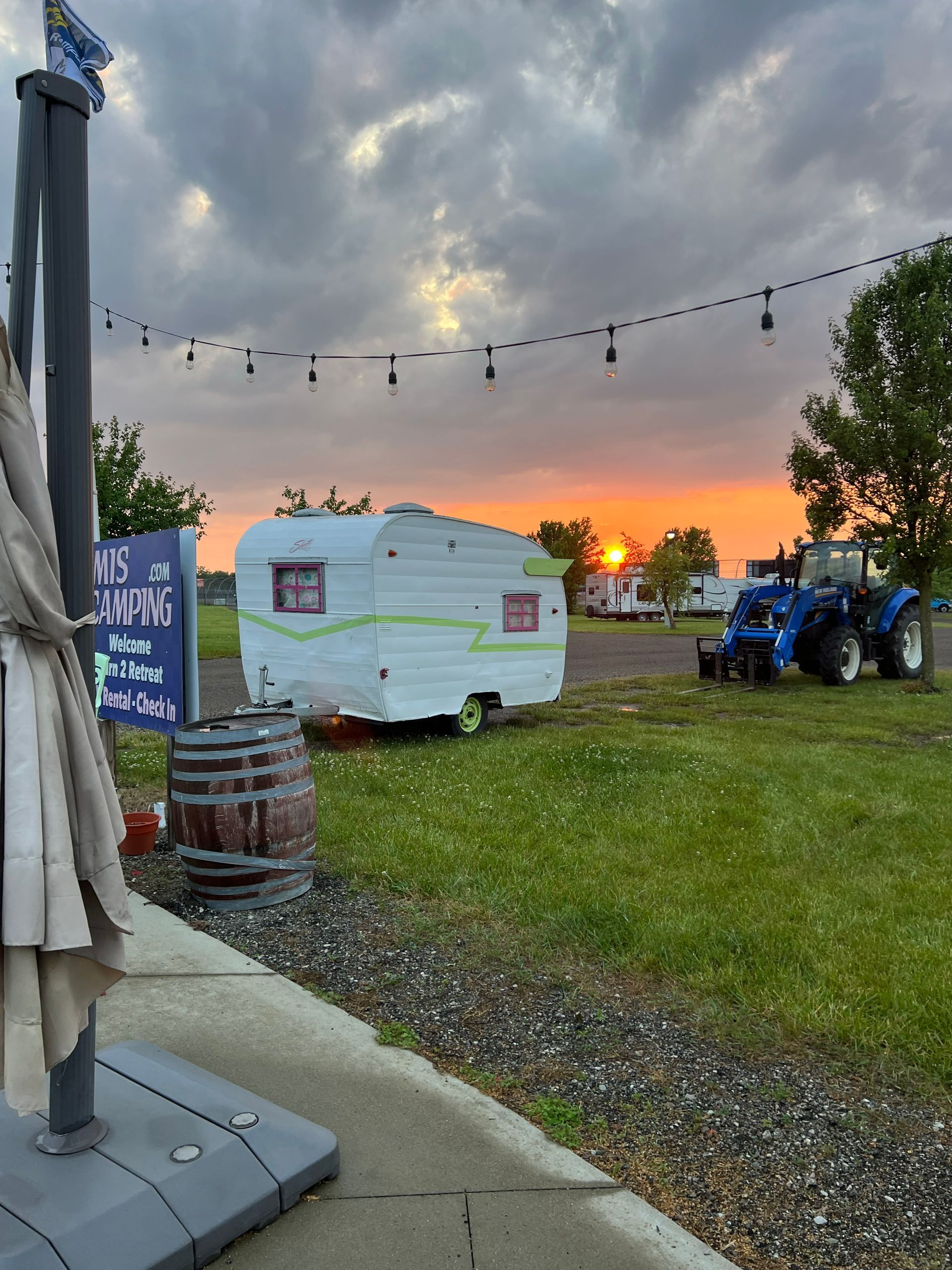 Sunset over a grassy field with a vintage camper, tractor, string lights, and a partially visible patio umbrella.