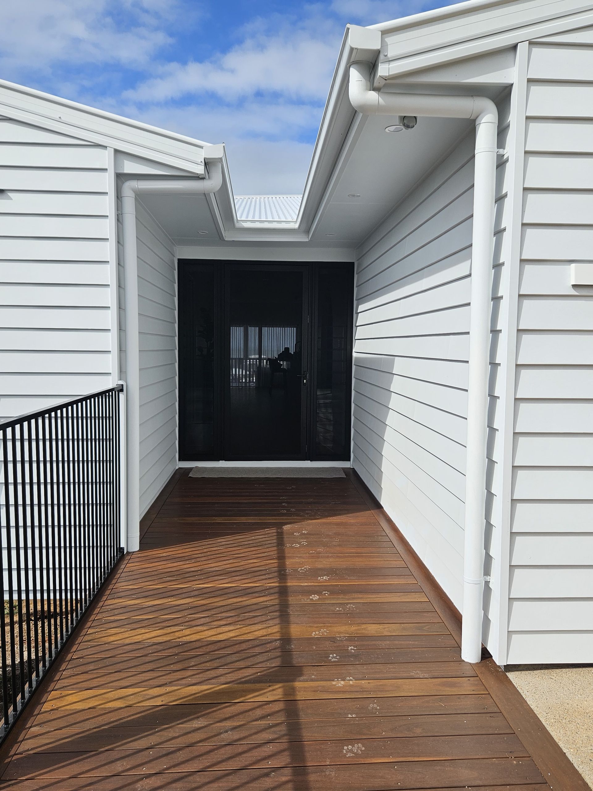 A wooden walkway leads to a dark glass front entrance, flanked by white horizontal-sided walls under a clear blue sky.