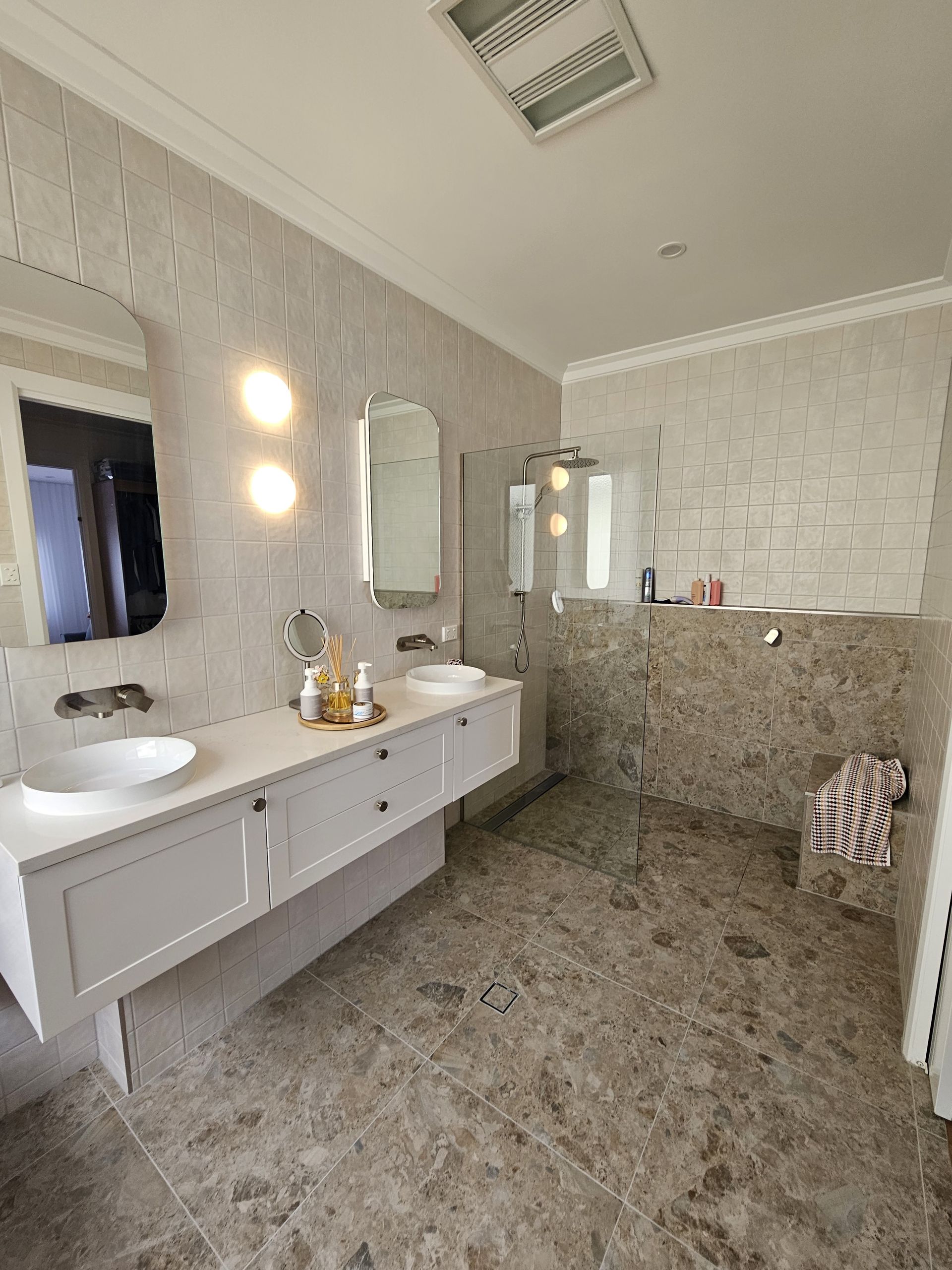 Modern bathroom with a white floating vanity, two round mirrors, and a walk-in glass shower with stone tile flooring — Jones Building Group in Yeppoon, QLD