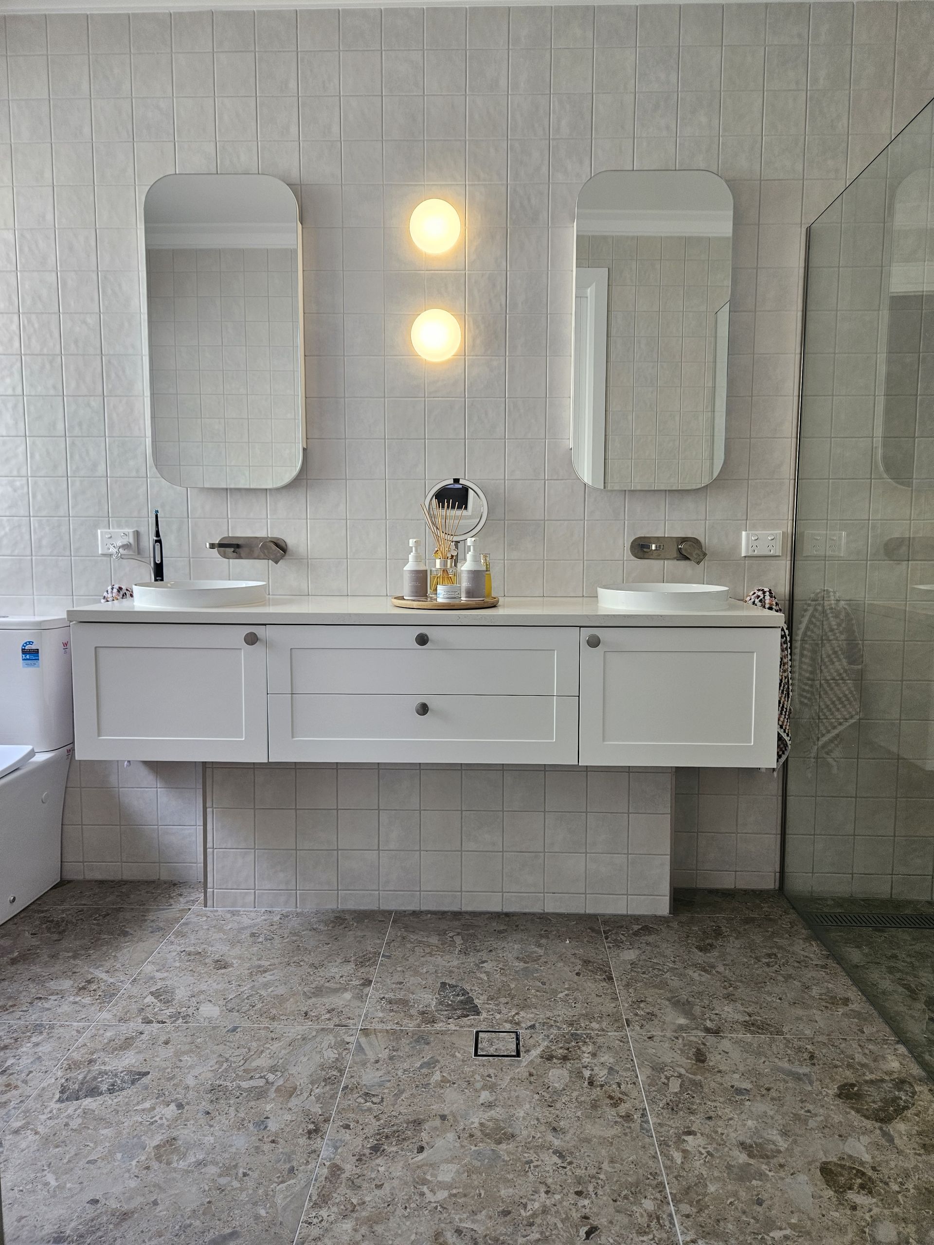 A modern double-sink bathroom vanity with white cabinetry, two rounded mirrors, warm lighting, and stone flooring.