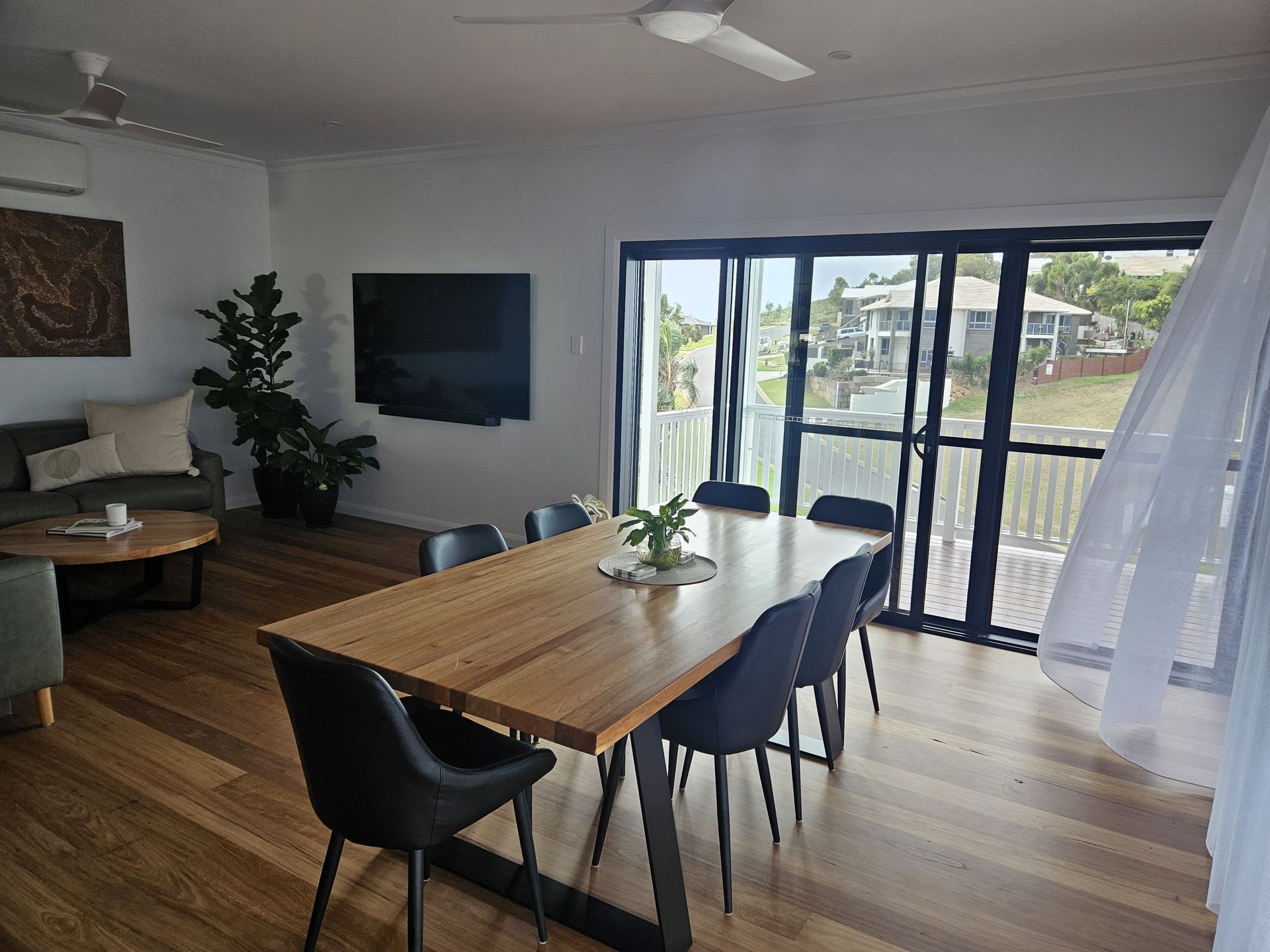 A bright living and dining room with a long wood dining table, black chairs, a TV, and glass doors to an outdoor view.