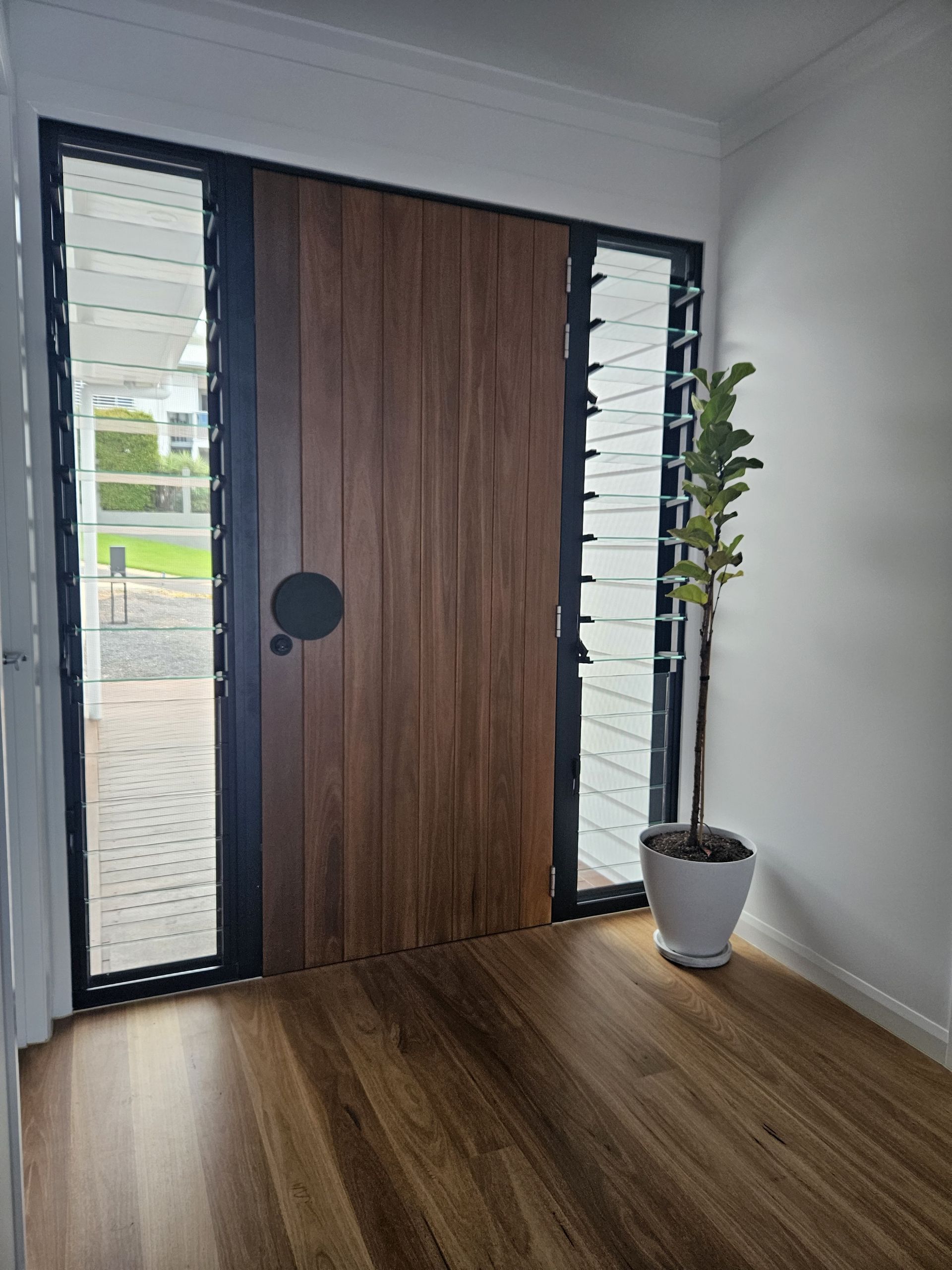 A wooden front door with a round black handle, flanked by tall louvered windows and a potted plant on a hardwood floor — Jones Building Group in Yeppoon, QLD