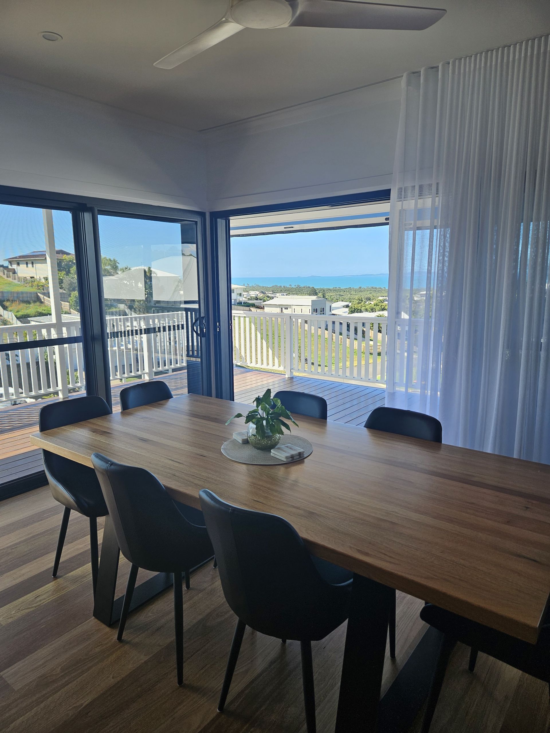 A modern wooden dining table with black chairs overlooks a sunny coastal landscape through large glass doors — Jones Building Group in Yeppoon, QLD