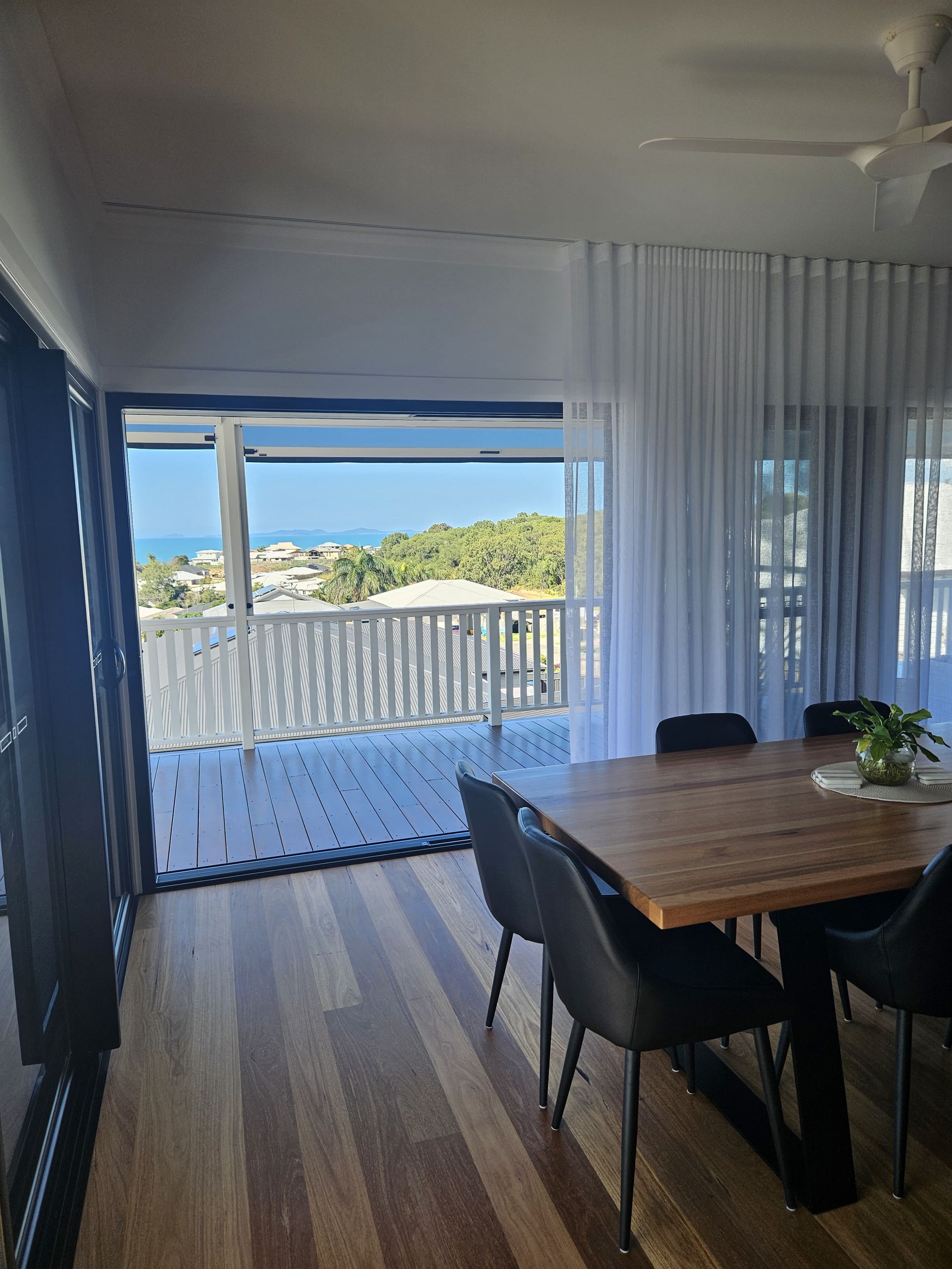 Dining area with a wooden table and black chairs, next to large sliding doors looking out onto a deck with an ocean view.