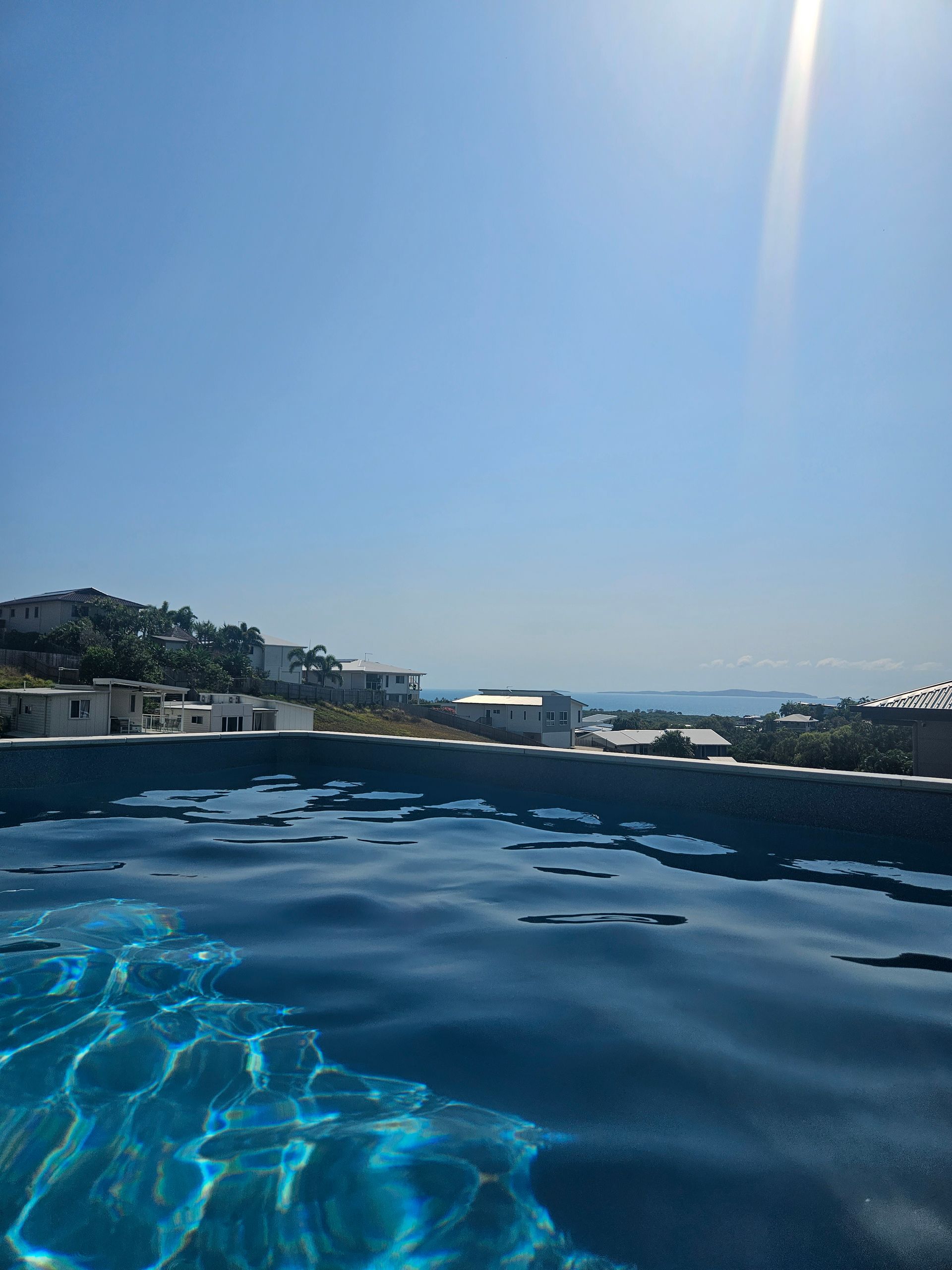 A bright sun shines over a dark blue swimming pool, with white buildings and distant islands visible on the horizon — Jones Building Group in Yeppoon, QLD