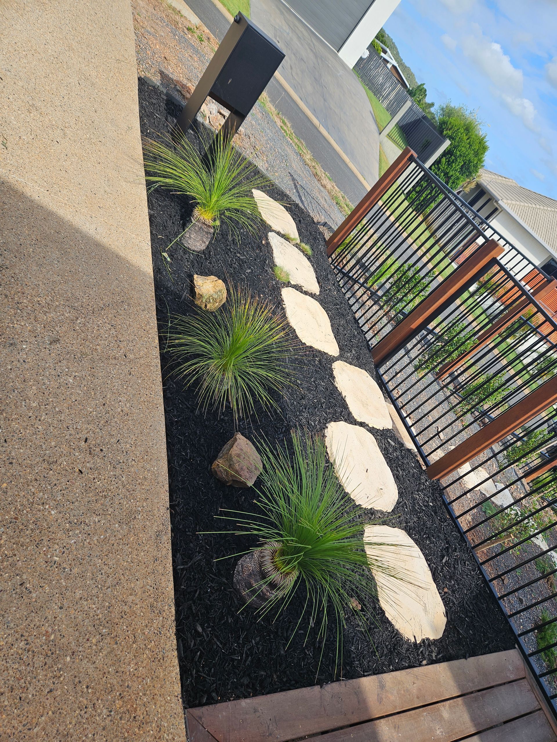 A landscaping strip with black mulch, stepping stones, small spiky plants, and decorative rocks beside a fence — Jones Building Group in Yeppoon, QLD