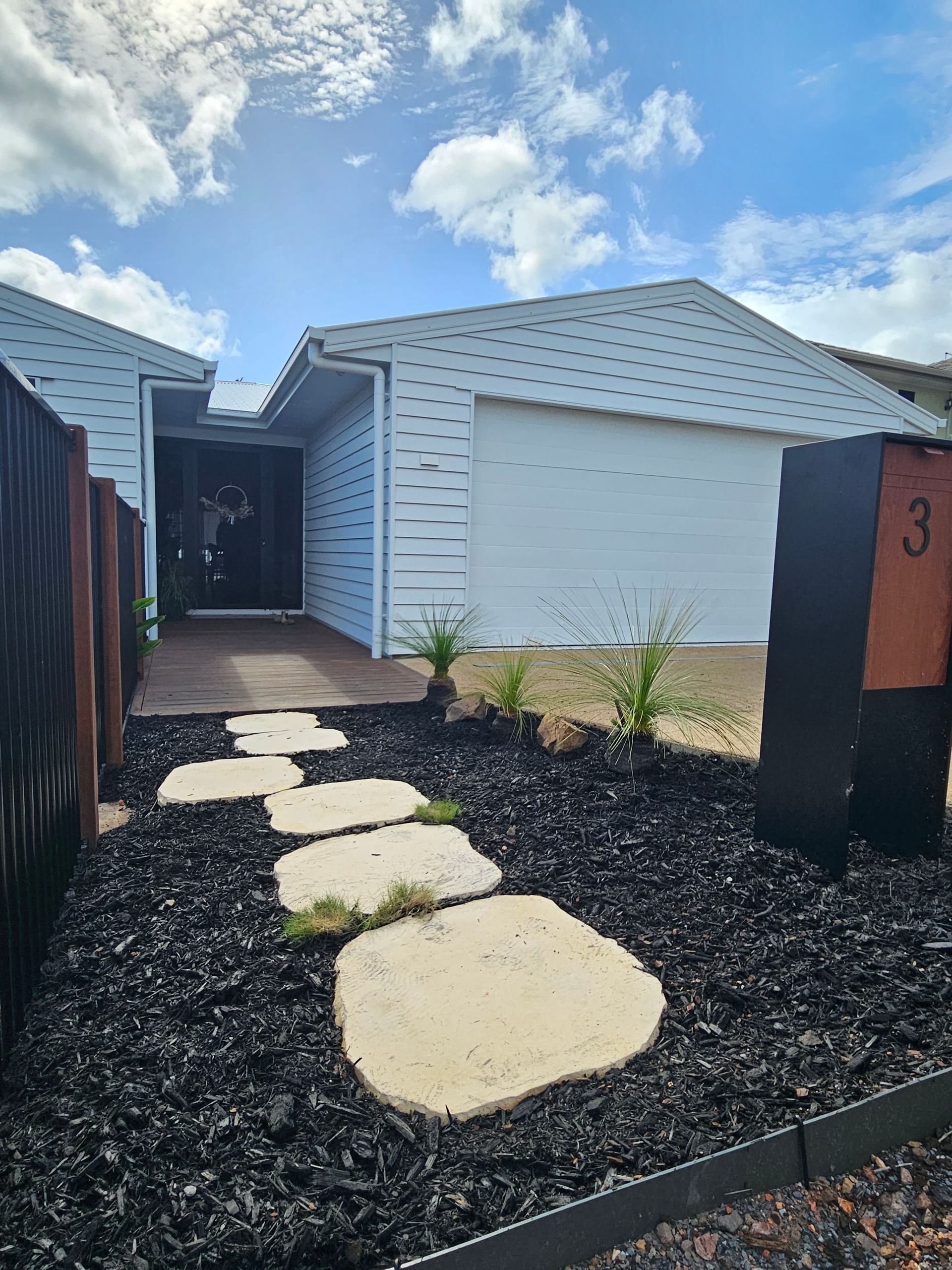 A modern home exterior with a white garage, black doorway, a stone path through black gravel, and a house number 3 sign — Jones Building Group in Yeppoon, QLD