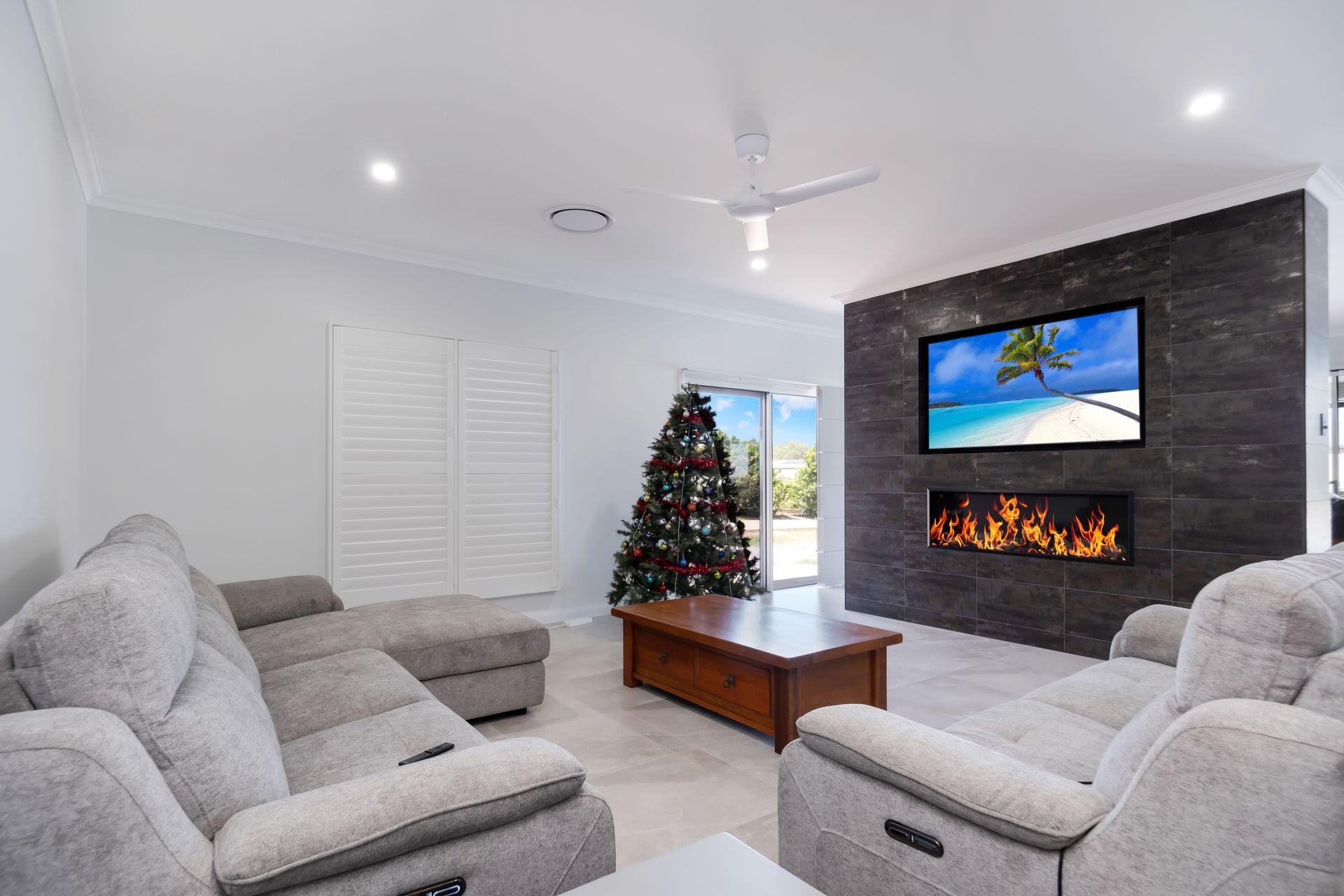 A living room with gray sectionals, a wooden coffee table, a Christmas tree, and a mounted TV above a linear fireplace.