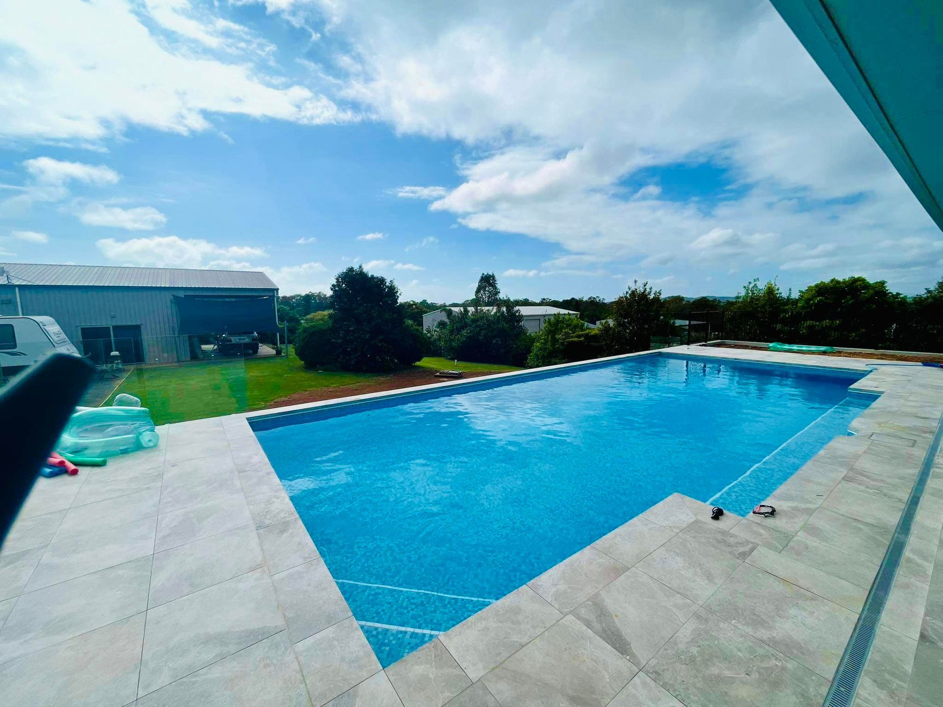 A large, bright blue rectangular swimming pool surrounded by grey stone patio tiles under a partly cloudy sky.
