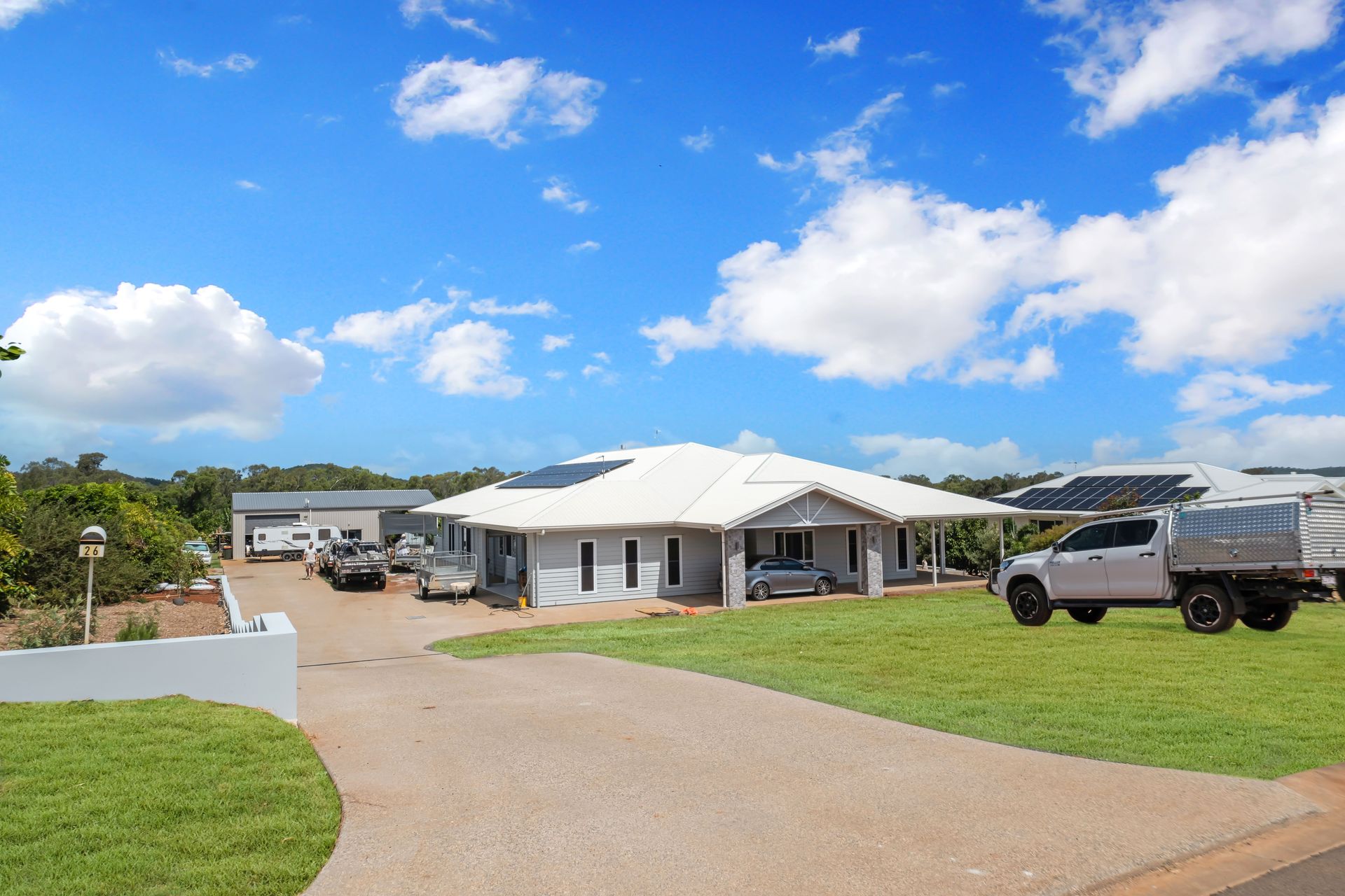 A modern light-gray house with a white roof and solar panels sits on a green lawn under a bright blue, cloudy sky — Jones Building Group in Yeppoon, QLD