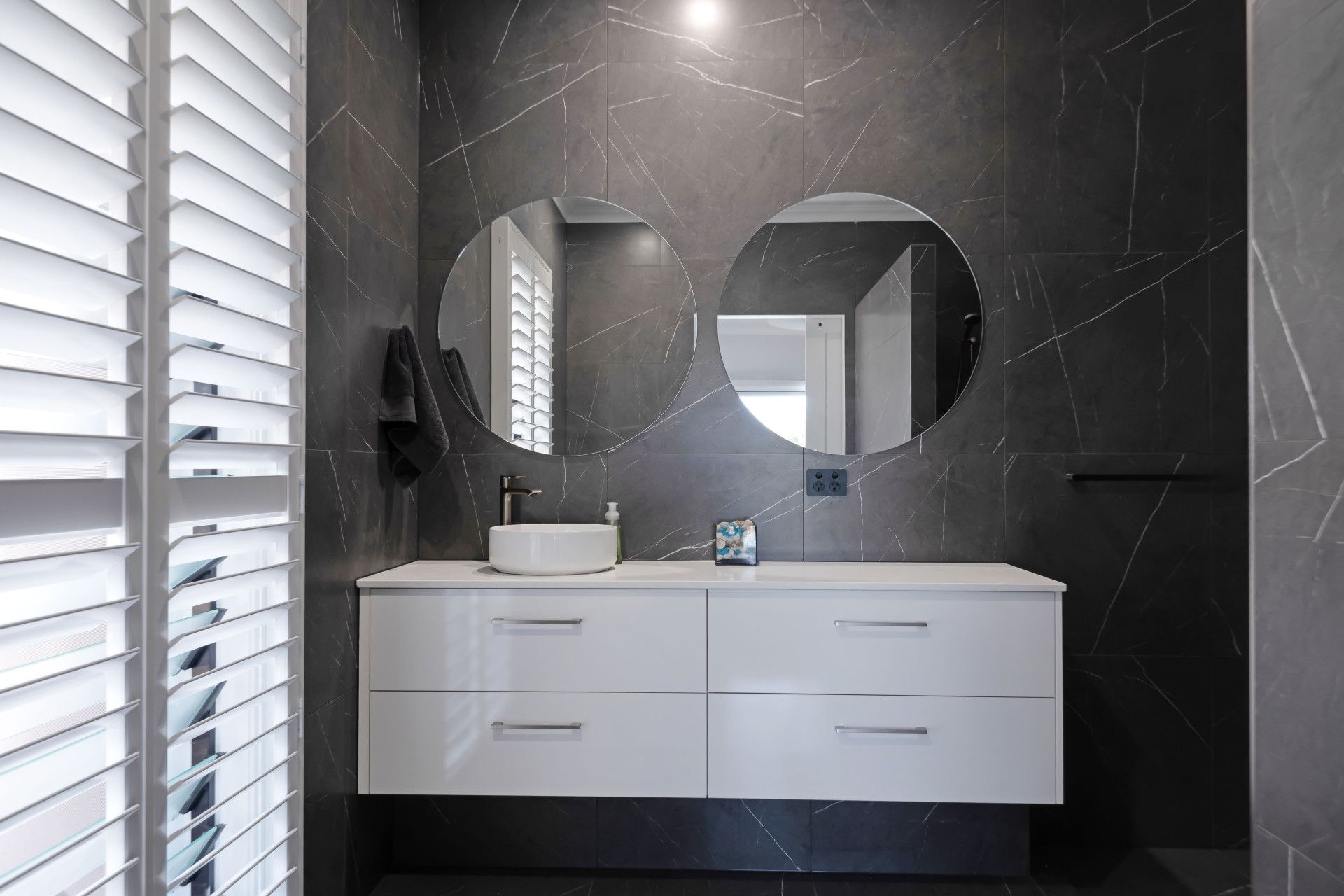Modern bathroom with a white vanity, a round vessel sink, two circular mirrors, and dark marble-look walls.