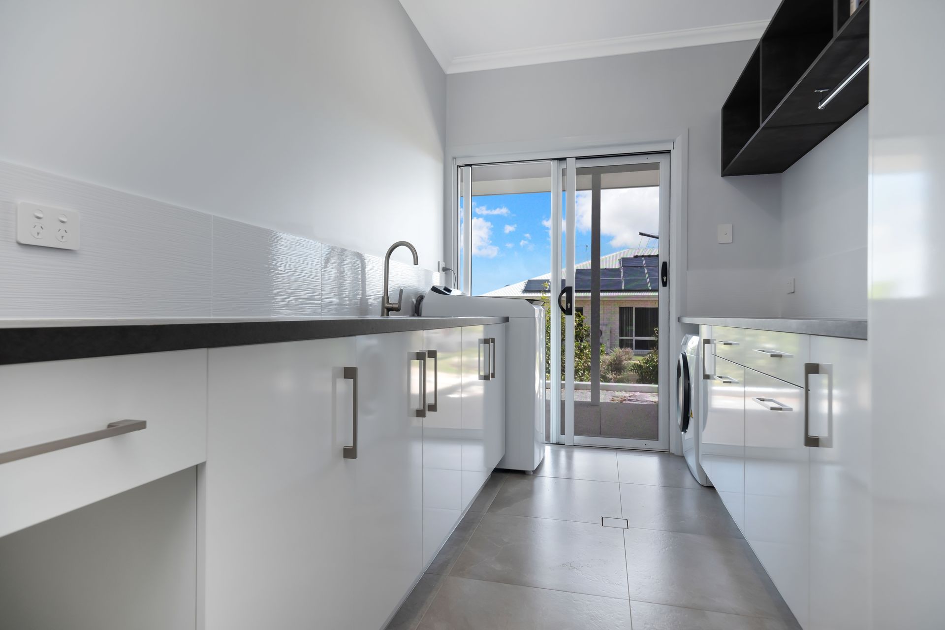 A clean, modern laundry room with white cabinets, dark countertops, a sink, a washing machine, and sliding glass doors — Jones Building Group in Yeppoon, QLD