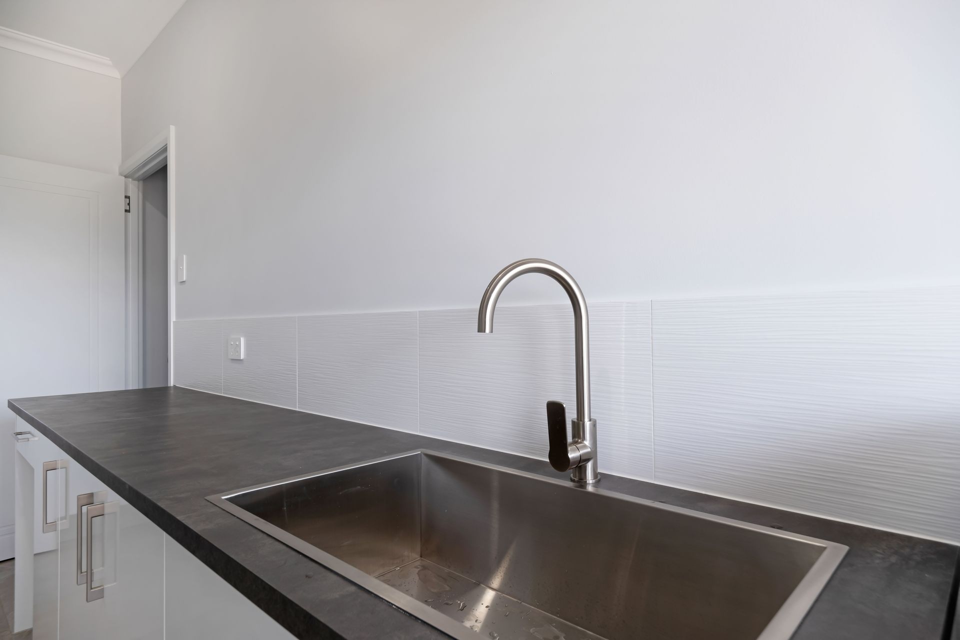 A stainless steel sink with a modern curved faucet set into a dark gray countertop against a white tiled backsplash — Jones Building Group in Yeppoon, QLD