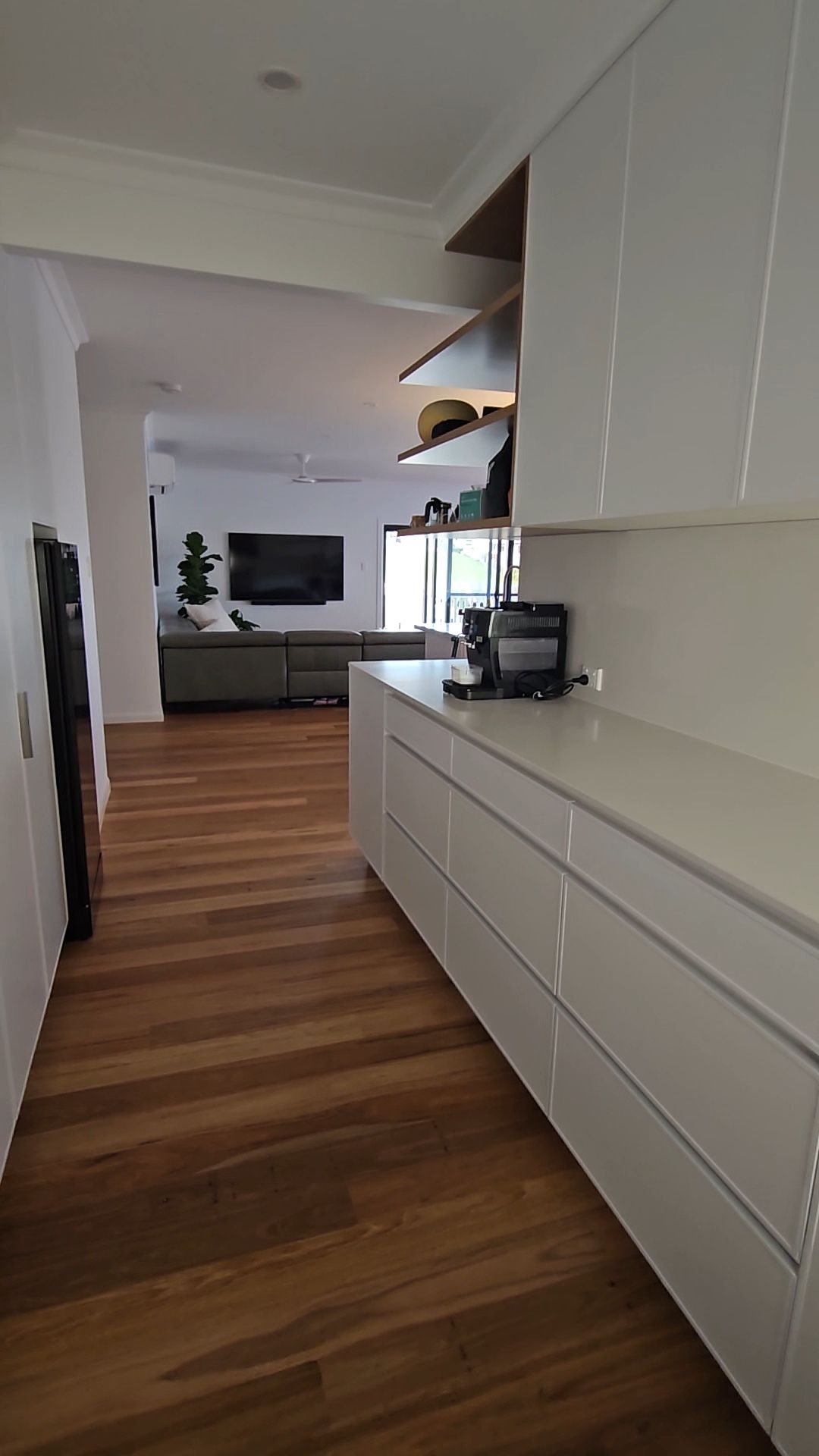 A bright kitchen interior with white cabinetry and wood flooring, leading into an open living room with a gray sofa — Jones Building Group in Yeppoon, QLD