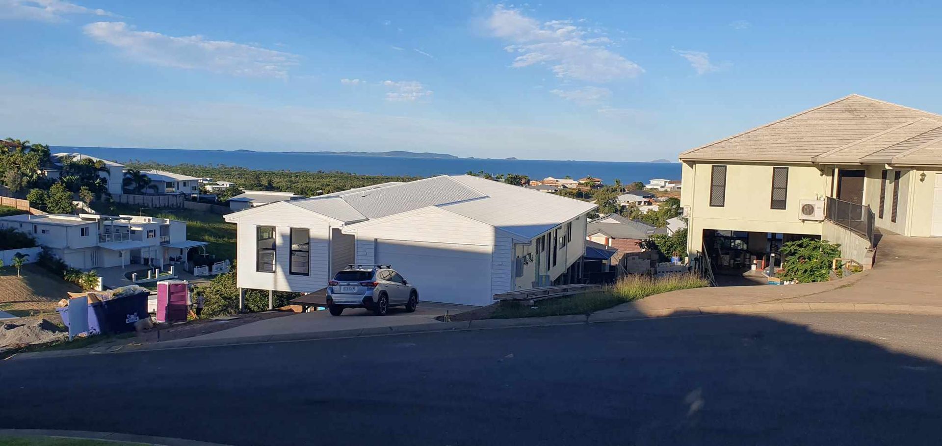 A suburban street with homes on a hillside overlooking a blue ocean under a clear, bright sky.