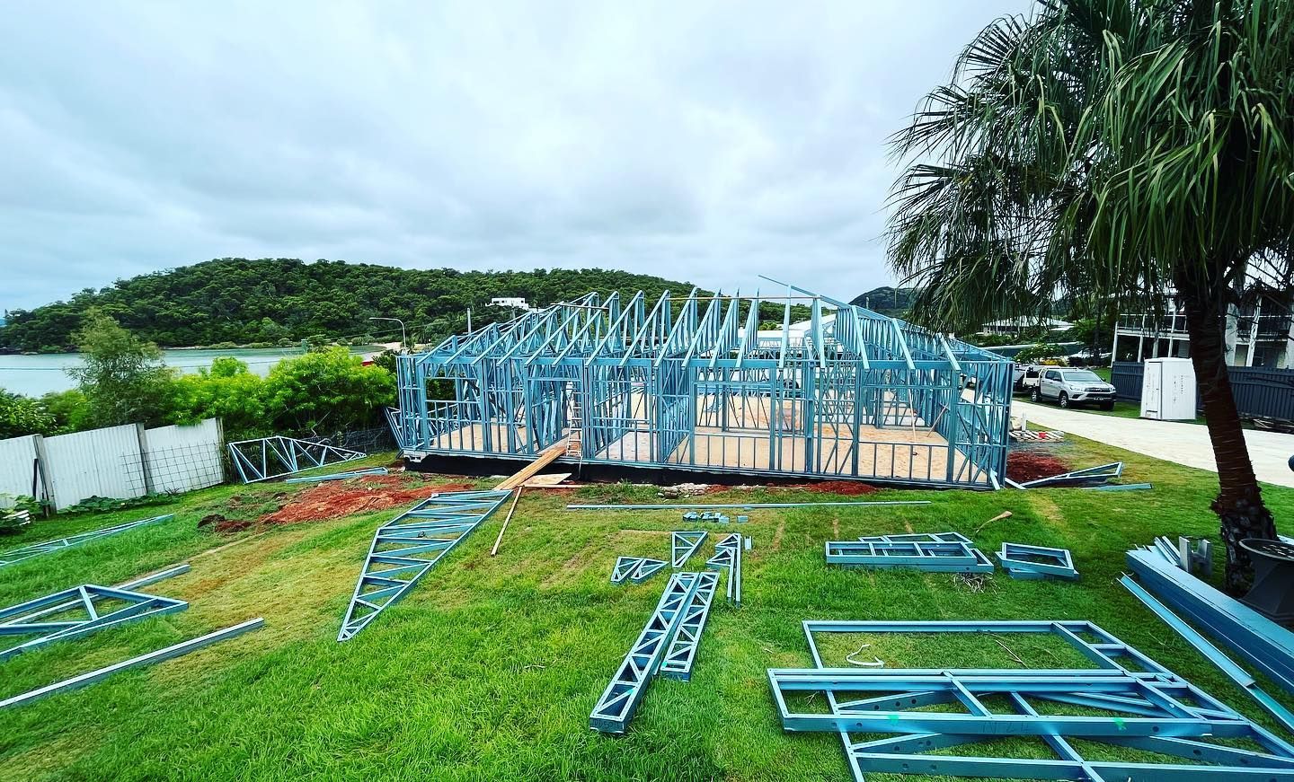 The metal frame of a house under construction sits on a grassy lot near a body of water, surrounded by building parts — Jones Building Group in Yeppoon, QLD