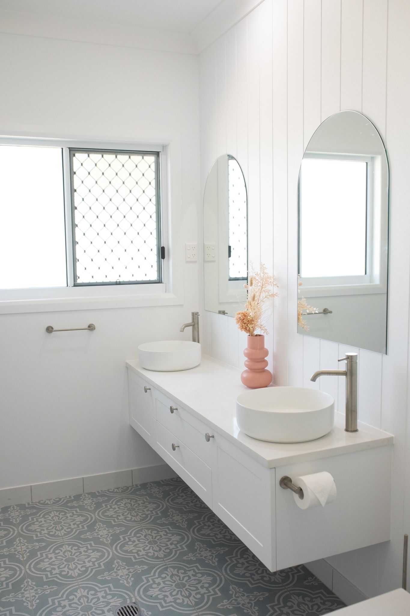 A modern white bathroom with a floating vanity, twin vessel sinks, arched mirrors, and patterned blue and white floor tiles — Jones Building Group in Yeppoon, QLD