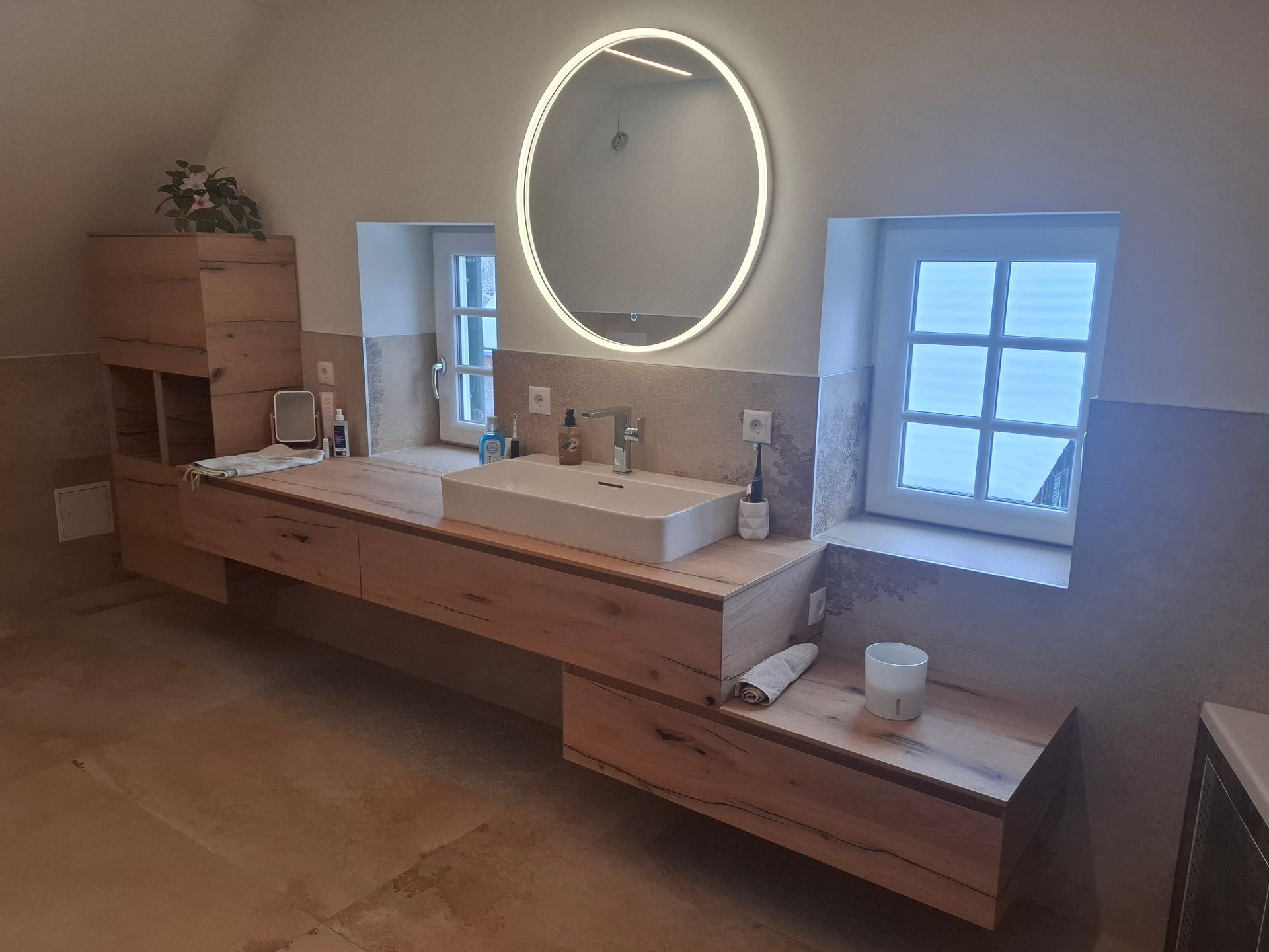 Badezimmer mit schwebendem Waschtisch aus Holz, großem beleuchtetem Spiegel und zwei kleinen Fenstern.