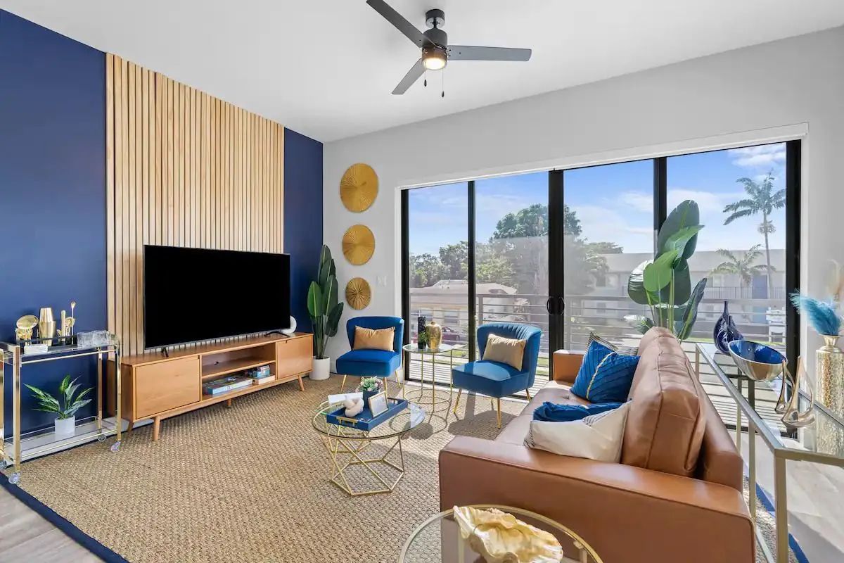 Bright modern living room with blue chairs, tan sofa, wall TV, and large windows overlooking palm trees.
