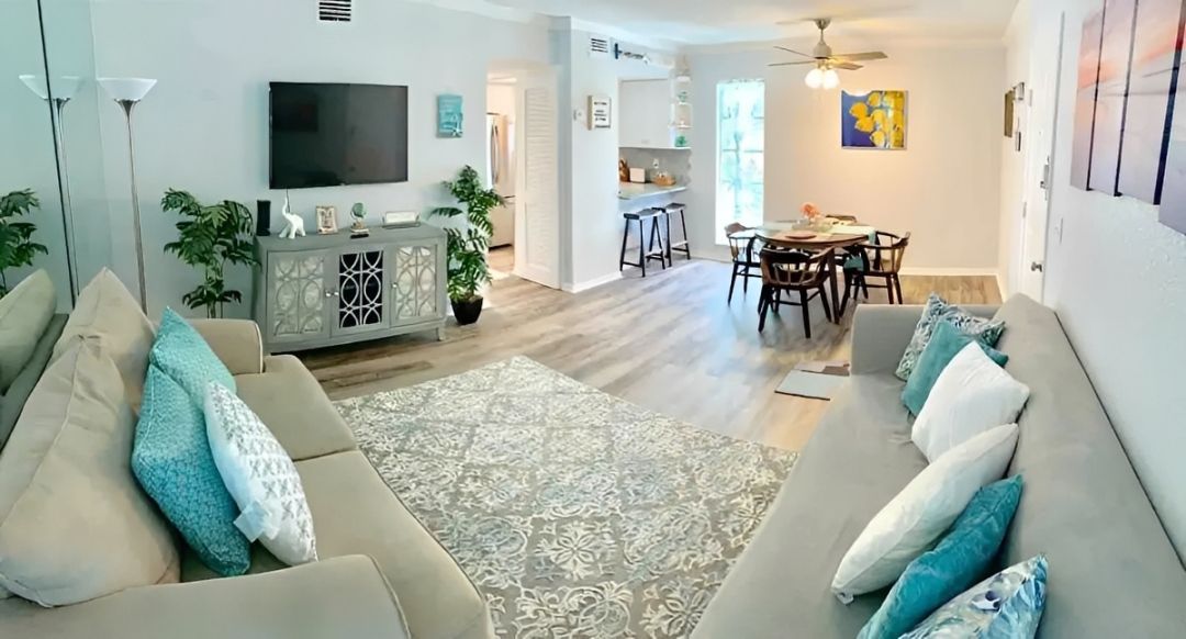 Bright living and dining room with gray sofas, teal pillows, TV, and patterned rug